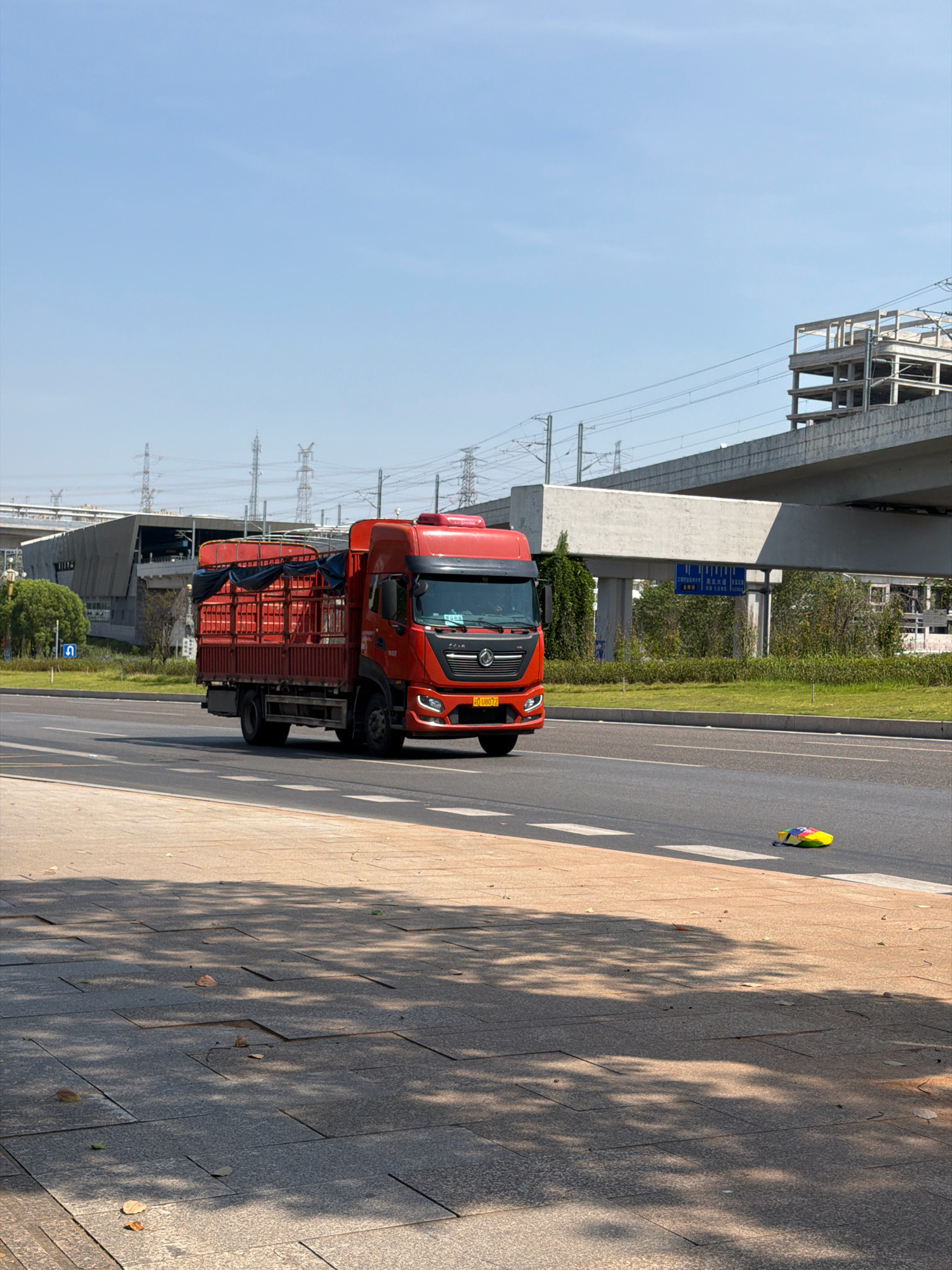 A bright red truck is captured in motion on a paved road, its bed filled with red crates covered by a dark tarp. The truck has a license plate that reads "HQ 08072". In the background, an elevated structure, possibly part of a transportation system, is under construction. Power lines and pylons are visible against a clear blue sky, indicating an urban or suburban setting. To the right of the road, a colorful object, perhaps a piece of luggage or a discarded item, lies on the ground. The foreground shows a textured sidewalk with scattered leaves, dappled with sunlight and shadow. The scene appears to be in Jiangjin, China, given the context provided by the user.
