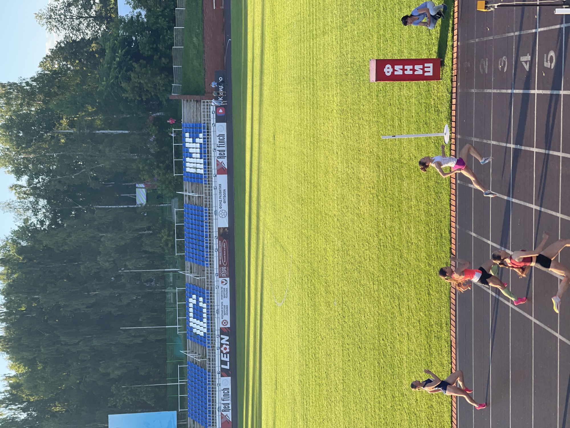 An outdoor athletic track is depicted, featuring a race in progress. Several female runners are seen on the track, one of whom is nearing the finish line, indicated by a red sign with white Cyrillic lettering that reads "ФИНИШ" (FINISH). Another runner is to the left, also in mid-stride. Further down the track, two other runners are visible. Spectators are present, with one woman crouching near the finish line and another in the stands in the background. The track is surrounded by a green grassy field, which is illuminated by sunlight, casting long shadows. In the background, there are stadium bleachers with blue seating, and beyond that, a dense line of trees under a clear sky. The overall scene suggests a competitive sporting event.