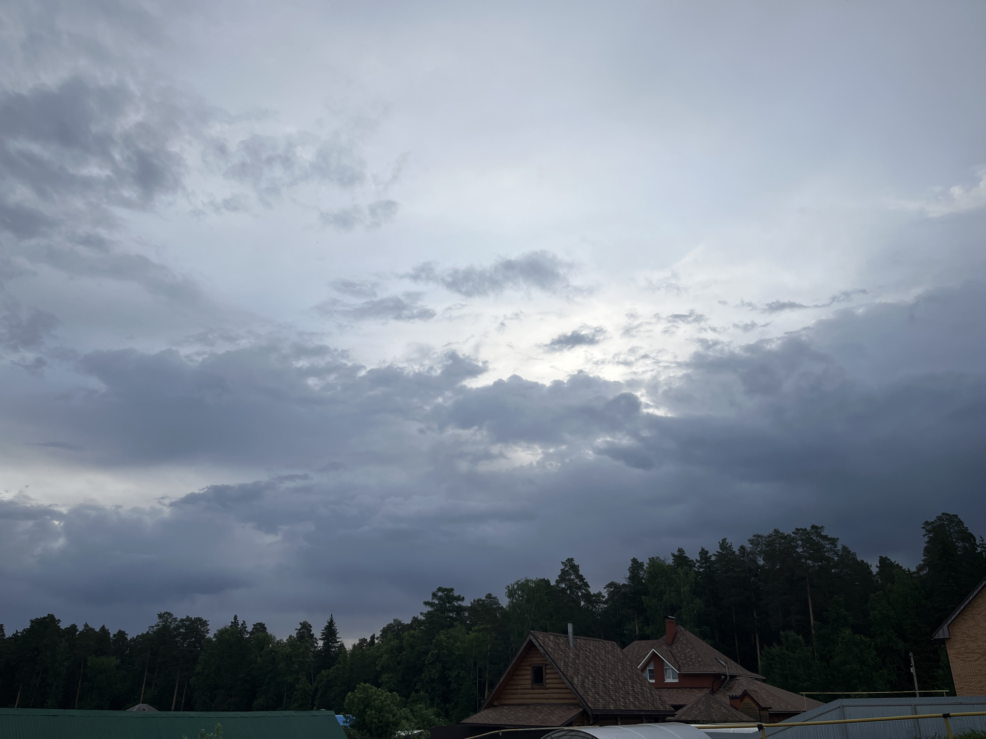 The image shows a landscape of Chaikovsky, Russia, under a cloudy sky. Dense trees form a line on the horizon. In the foreground, there are buildings, including a house with a brown roof, some with greenhouses attached. The sky is overcast with dark gray clouds.