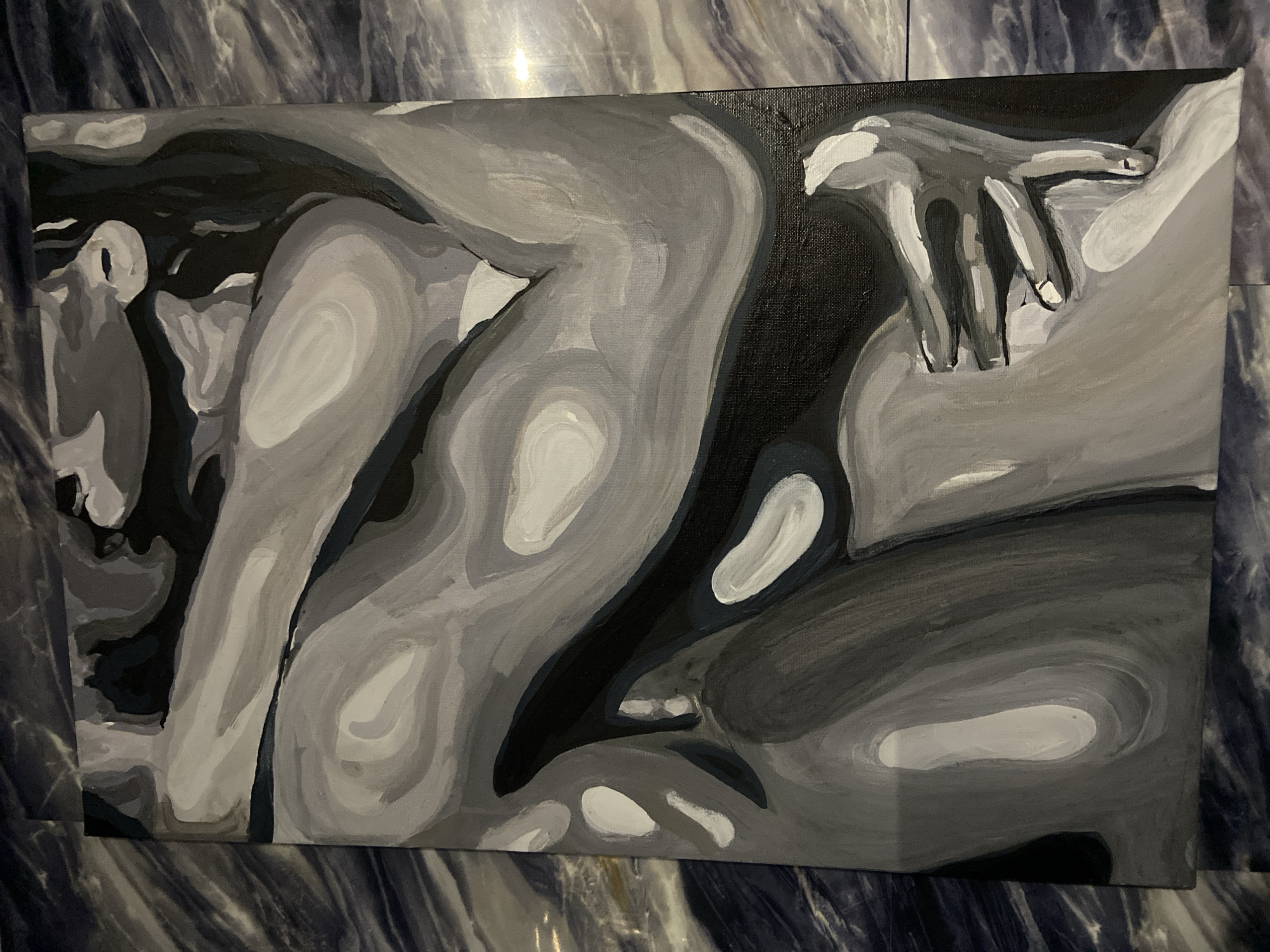 The image is a close-up, vertical shot of a grayscale painting on a canvas. The painting depicts an abstract, intertwined embrace of two figures, rendered in various shades of gray and black, with highlights of white. The forms are fluid and suggest curves of the human body. The canvas is placed on a surface with a dark, marbled pattern, which appears to be a floor or tabletop. The lighting casts subtle shadows and reflections on the painting and its surroundings. There is no discernible location within the city of Stavropol, Russia, or any other specific geographical context visible in the image.