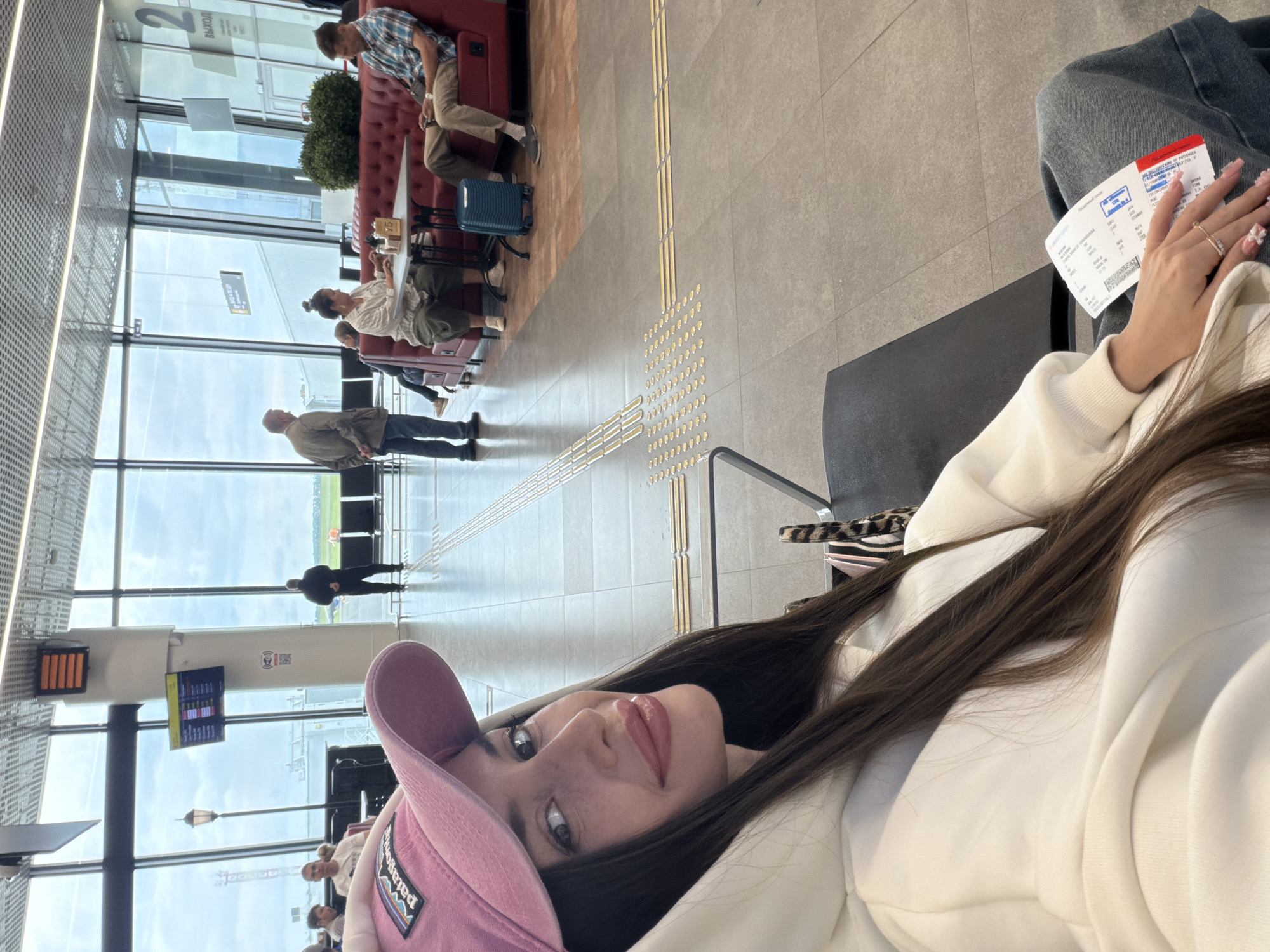The image captures a selfie taken by a young woman in what appears to be an airport waiting area, possibly in Новое Мартьяново, Russia. The woman is positioned in the foreground, lying or leaning back, looking towards the camera. She has long dark hair, is wearing a white hoodie, and a pink baseball cap with the "Patagonia" logo visible. Her left hand, adorned with rings and manicured nails, holds a boarding pass displaying text such as "ПОСАДОЧНЫЙ ТАЛОН" (Boarding Pass), "PASSEGER", "BOARDING TIME", "DATE", "FLIGHT", "GATE", "SEAT", "CLASS", "ПРИБЫТИЕ" (Arrival), and "ОТПРАВЛЕНИЕ" (Departure).
The setting features large windows in the background, revealing a cloudy sky and an airfield with distant figures, indicating daylight hours. The interior floor is tiled and includes tactile paving with raised dots and lines, designed for visually impaired people. In the mid-ground, several other travelers are visible. A man and a woman are seated at a table with red upholstered booths, and a blue carry-on suitcase rests beside them. Another man stands near the windows. A sign in the distance clearly displays "ВЫХОД" (Exit) in Cyrillic. Display screens, likely showing flight information, are also visible. The overall scene depicts a calm waiting period at an airport.