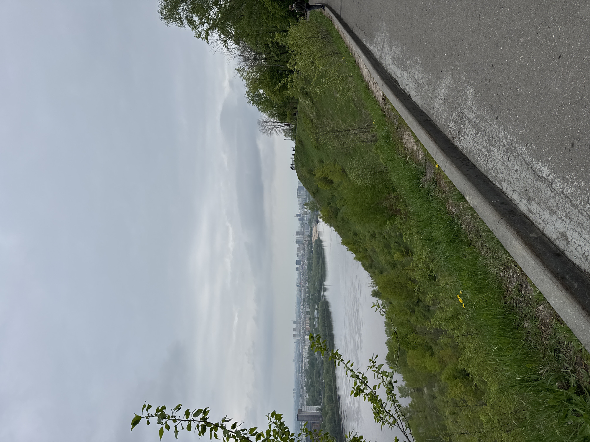 From an elevated perspective along a roadway in Nizhny Novgorod, Russia, a panoramic view unfolds. The scene features a river winding through the urban landscape, flanked by dense greenery on its banks. Buildings of varying heights punctuate the skyline in the distance, suggesting a developed urban area. Overcast skies create a muted, gray backdrop. A few people can be spotted along the elevated area, observing the vista. The foreground is dominated by grass and some trees with foliage, growing on the slope. A concrete road edge is visible.