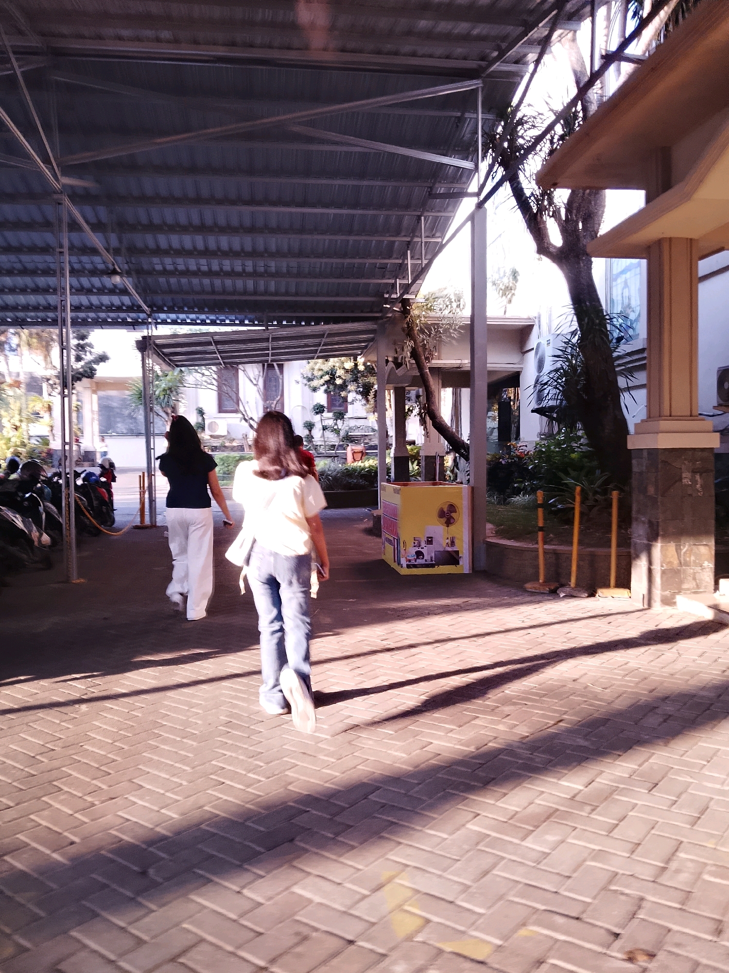 The image shows two women walking away from the camera on a brick-paved pathway in what appears to be an outdoor corridor or covered walkway. The woman in the foreground wears a light-colored short-sleeved top and blue jeans, while the woman behind her wears a dark top and white trousers. Sunlight casts long shadows across the brickwork.
To the left of the pathway, motorcycles are parked under a metal roof structure. In the midground, to the right, a yellow vending cart with graphics is visible, accompanied by several wooden posts. Beyond the cart, there is greenery and a portion of a building with a pillar. The building has a light-colored facade and some visible air conditioning units. The overall setting suggests a public space, possibly a market, a school, or a commercial area.
Based on the architectural style, vegetation, and the presence of a vending cart commonly seen in Indonesia, this scene is likely located in Malang, Indonesia. The atmosphere is bright, with direct sunlight indicating daytime.