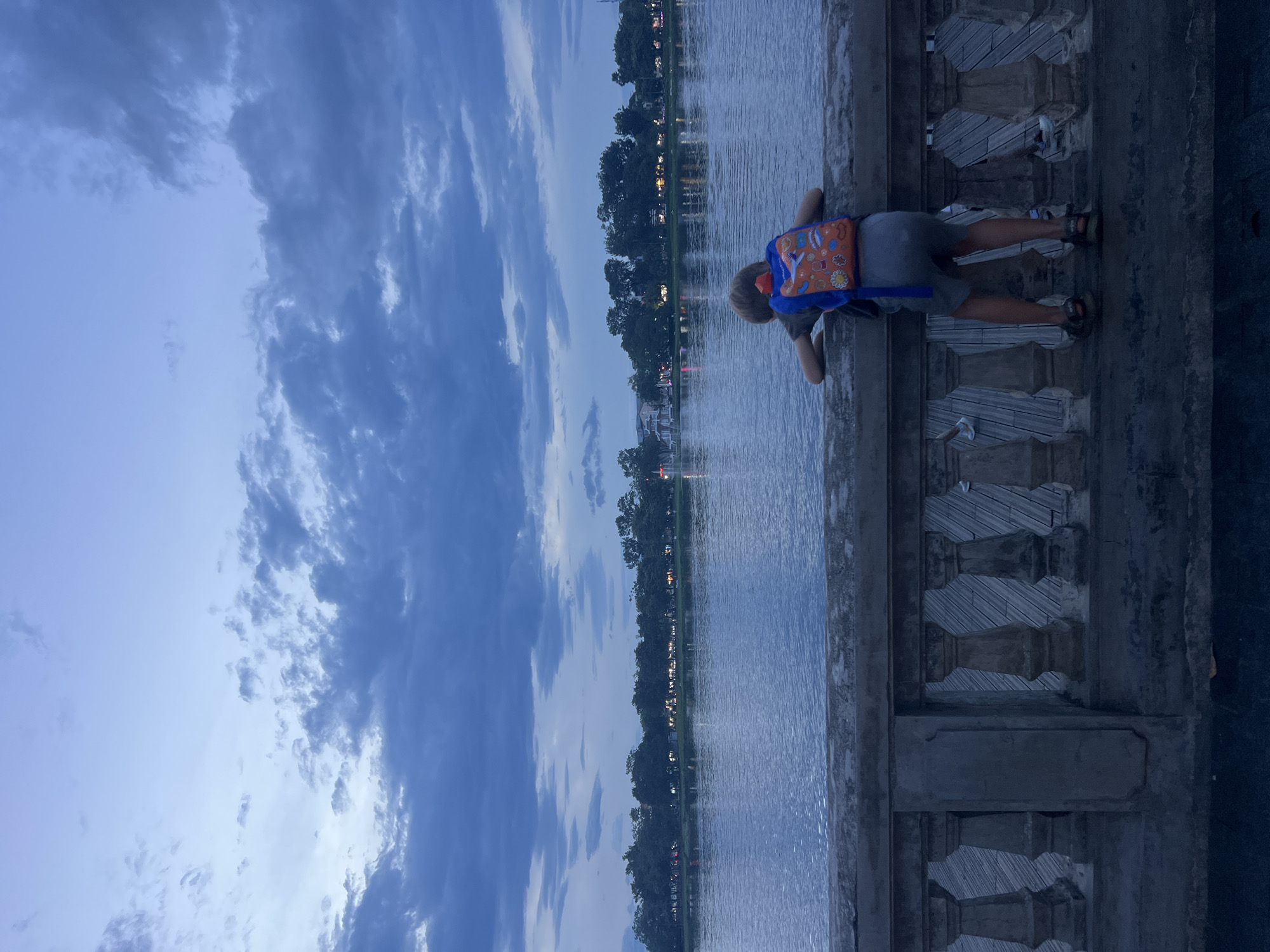 A young boy, seen from behind, leans over a stone railing, looking out at a body of water. He wears a gray t-shirt, gray shorts, and a colorful backpack. The railing is ornate with balusters and a rough-hewn stone top. Wooden planks are visible beneath the railing. Across the water, a tree line with buildings and lights can be seen. The sky above is cloudy and a deep blue, suggesting it is either early morning or late evening. The scene takes place outdoors, near a waterfront, possibly a park or promenade. There is no indication this image is located in Thuận Hóa Ward, Vietnam.