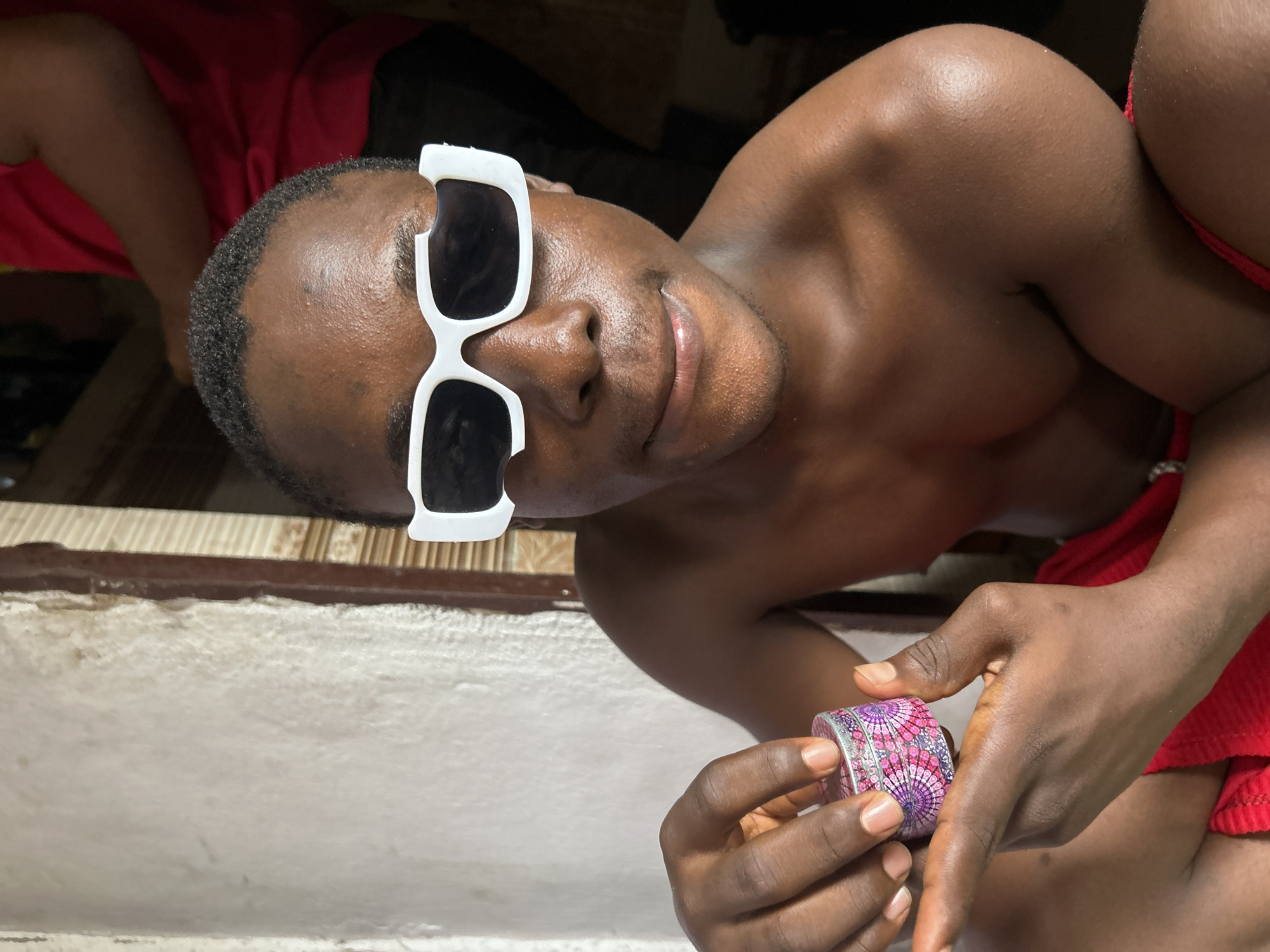In the city of Ihiagwa, Nigeria, a young Black male is captured in a close-up shot, seated outdoors. He is shirtless, wearing bright red shorts, and sporting a pair of white, avant-garde sunglasses that are notably broken on the left lens. His dark hair is short and textured. He is holding a small, round, metallic object with an intricate pink and purple geometric pattern, presenting it towards the camera with his left hand. The background is somewhat blurred, showing a textured white wall on the left and a doorway with vertical blinds on the right. Another person, wearing a red shirt, is visible in the upper right portion of the frame, also blurred. The lighting suggests it is daytime, with sunlight casting shadows and highlighting the sheen on the subject's skin.