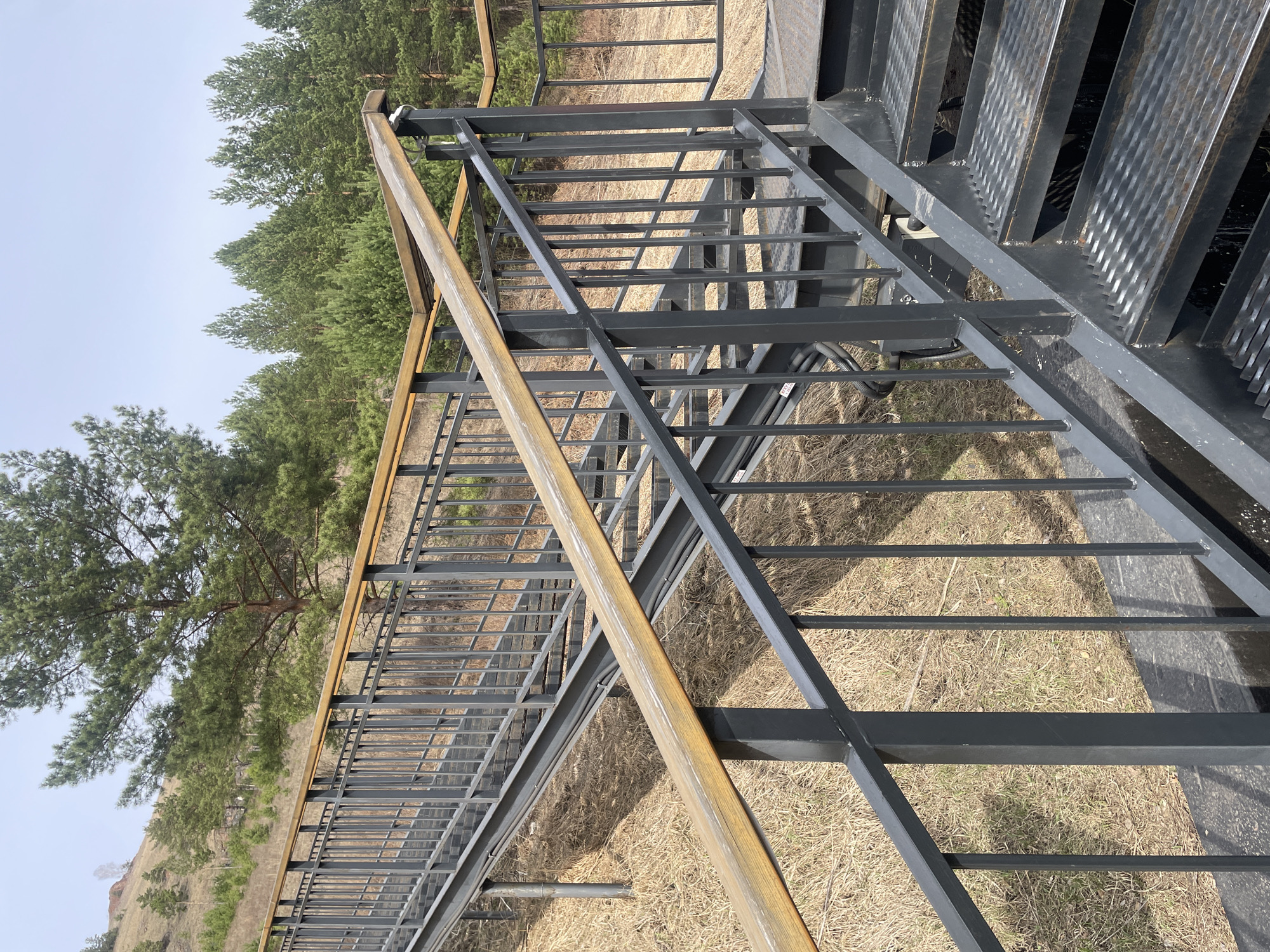 The image shows a metal staircase with a wooden handrail in an outdoor setting. The stairs are gray and have a textured surface. The handrail is light brown. The ground is covered in dry grass and foliage. In the background, there are trees and a hill under a sky. Based on the image analysis, the location may be in Krasnoyarsk, Russia.