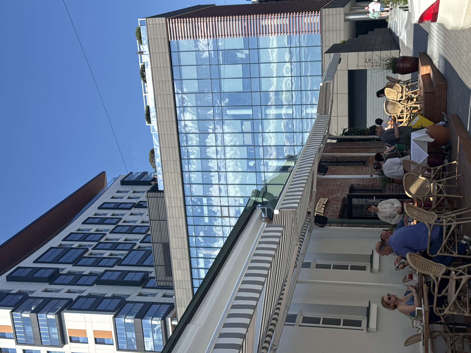 The image depicts an outdoor seating area of a restaurant in Yekaterinburg, Russia, with a modern multi-story building in the background. Several patrons are seated at tables under striped awnings. A woman in a blue top and sunglasses is at a table on the left, and a man in a blue shirt is seated opposite her, looking at his phone. Other diners are visible at tables further back. The restaurant appears to be called "ТОЛСТЫХ" (Tolstykh). The modern building features large glass windows reflecting the sky and clouds. A pathway with stairs leads up to the entrance of the modern building. People are walking on the pathway in the background. The day is sunny with a clear blue sky.