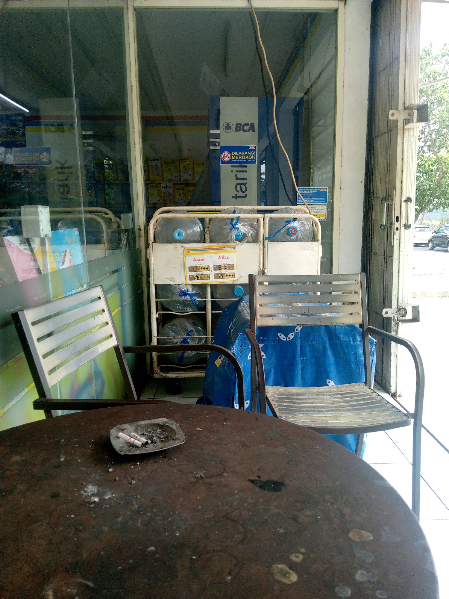 The image captures a view of a small storefront or minimarket in Cikupa, Indonesia, likely a convenience store. In the foreground, a round, weathered brown table is visible, with an ashtray containing two cigarette butts and ashes. Two metal chairs with slatted seats are positioned around the table. Behind the table and chairs, a metal rack holds several large gallon jugs of drinking water, some of which are covered with blue plastic. A sign above the water jugs displays prices for "Aqua" and "Oleo" brands. Through the glass storefront, an ATM machine with the "BCA" logo and a "dilarang merokok" (no smoking) sign are partially visible. The overall environment appears to be an outdoor area adjacent to a building with a metal security gate. The lighting suggests it is daytime, with bright sunlight illuminating the scene.