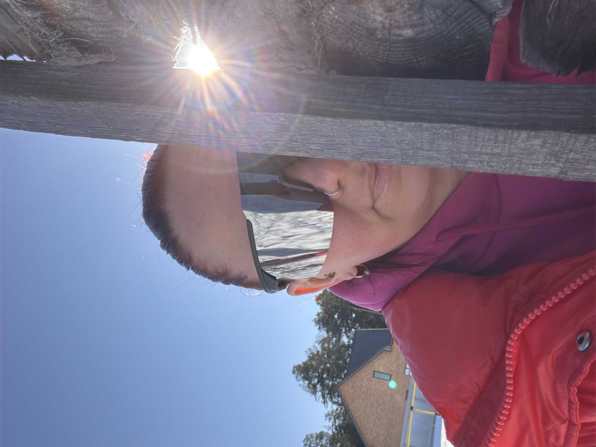The image shows a person wearing mirrored sunglasses, a pink hoodie, and a red jacket. The person is standing near a wooden structure, possibly a fence or building component. The sun is shining brightly, creating lens flares and reflecting off the sunglasses. The background includes a blue sky and a building with a light brown exterior and dark roof, as well as some trees. The location is potentially Chaikovsky, Russia, based on the implied association and the provided instructions.