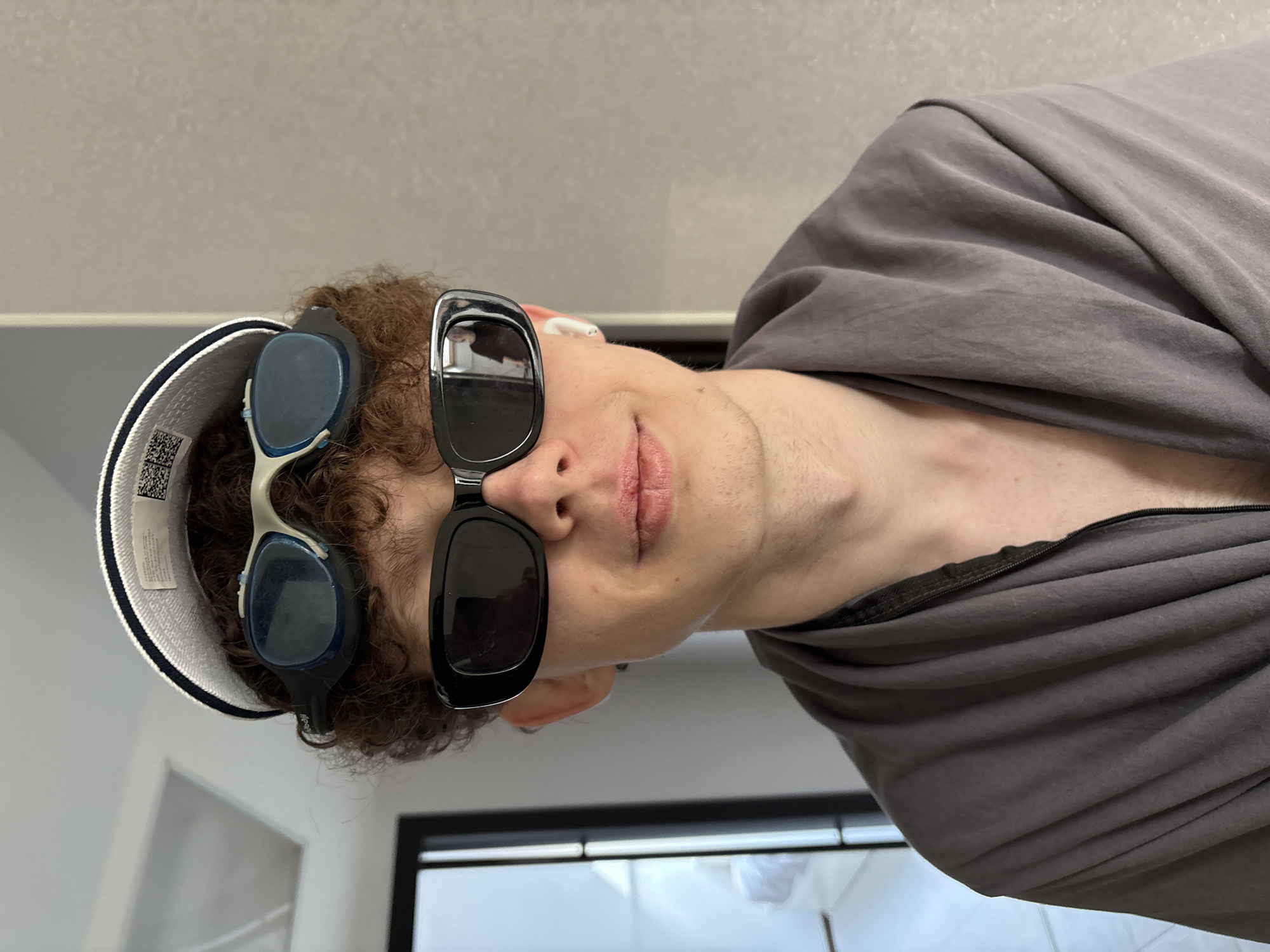 A person is depicted from a low, tilted angle. The individual wears sunglasses, with additional eyewear perched atop their head along with a cap. The person is dressed in a gray hooded garment. The background consists of a white wall, a window, and white tiled flooring. The image doesn't contain information to determine the location.