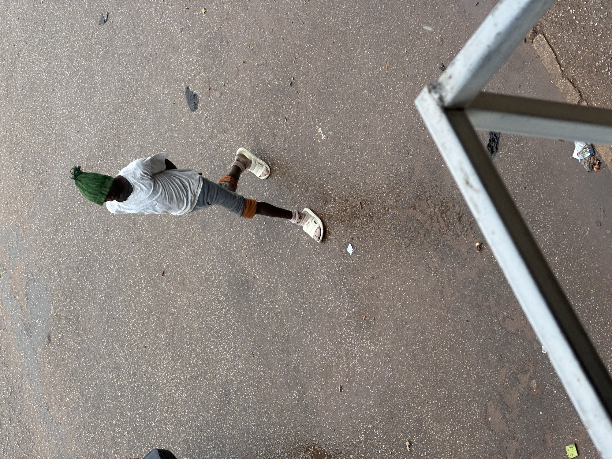A young boy wearing a green knitted hat, a light-colored t-shirt, and shorts walks on a wet asphalt road in Bukuru, Nigeria. He is wearing white sandals and socks. The road appears to be a street, possibly with some litter and debris scattered on it. The perspective is from above, looking down at the boy as he walks. The boy is captured in motion, with his left leg forward.