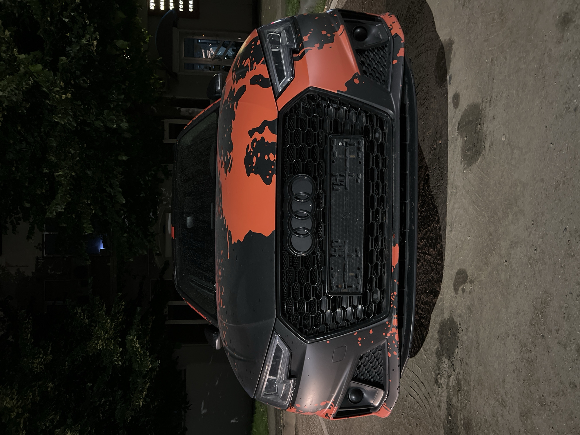 A frontal, eye-level medium shot shows the front of a dark grey Audi RS5 with an orange and black splatter paint job parked at night on a paved surface. The car's headlights are on, illuminating the grille and front bumper. The grille is black with a honeycomb pattern and features the Audi logo. A license plate holder is mounted in the center of the grille. Orange paint splatters are visible on the hood, bumper, and fenders of the car. In the background, dark green trees with some white flowers overhang a building with windows. To the right of the car, a sign with Russian text is partially visible, appearing to be for a restaurant or shop. The ground in front of the car is wet, with some dark stains. The scene appears to be in an urban environment, likely a street or parking lot. There are no people visible in the image.