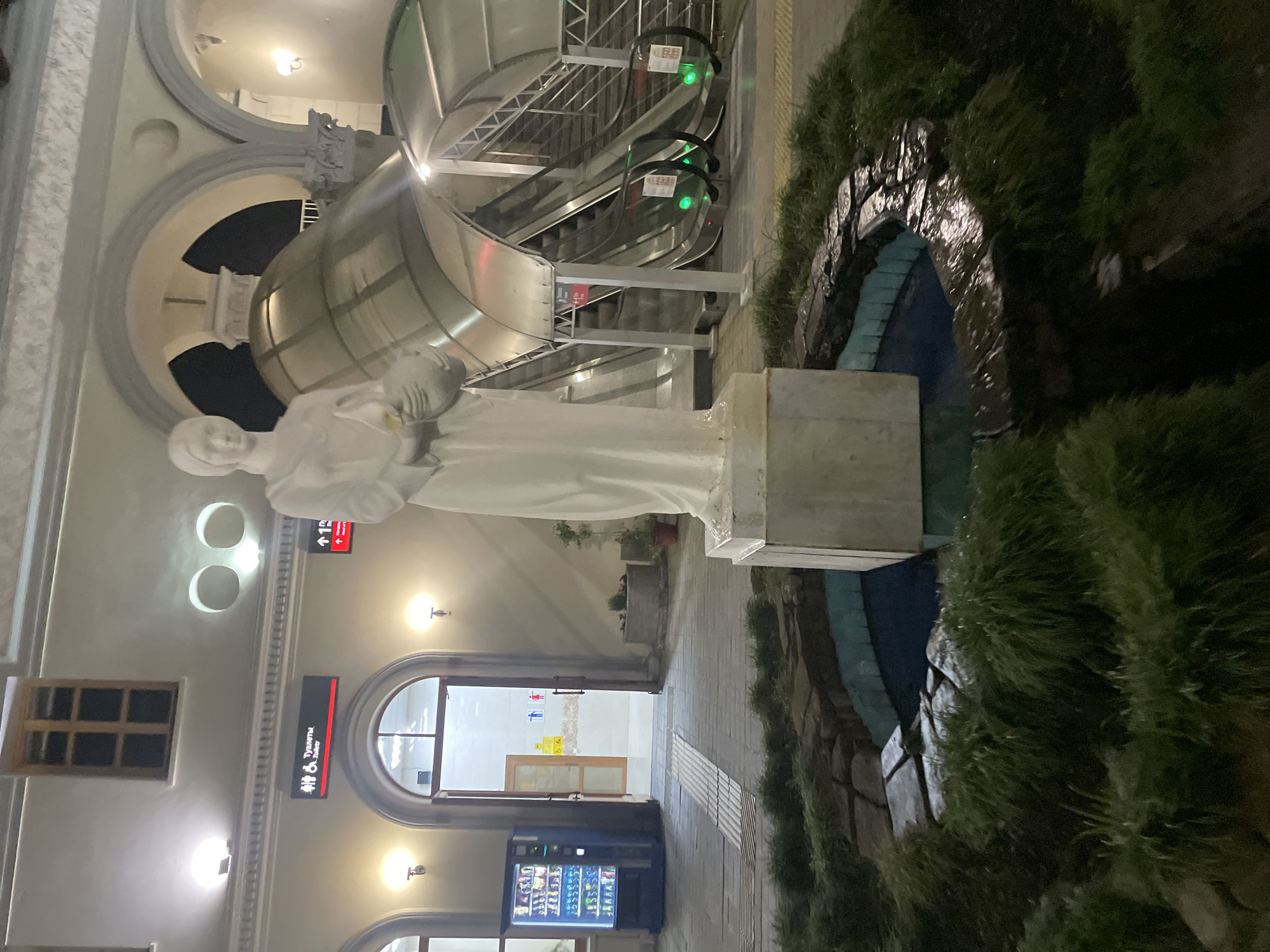 This image depicts the exterior of a building in Sochi, Russia, at night, featuring a statue and escalators. In the foreground, a white statue of a woman holding a vessel stands in a circular fountain surrounded by lush green grass and rocks. To the right, a covered escalator with a metallic, curved roof leads upwards. The building facade features arches, windows, and illuminated signs, including one indicating "Toilets." A vending machine is visible to the left. The overall scene suggests a public space like a train station or an entertainment venue.
