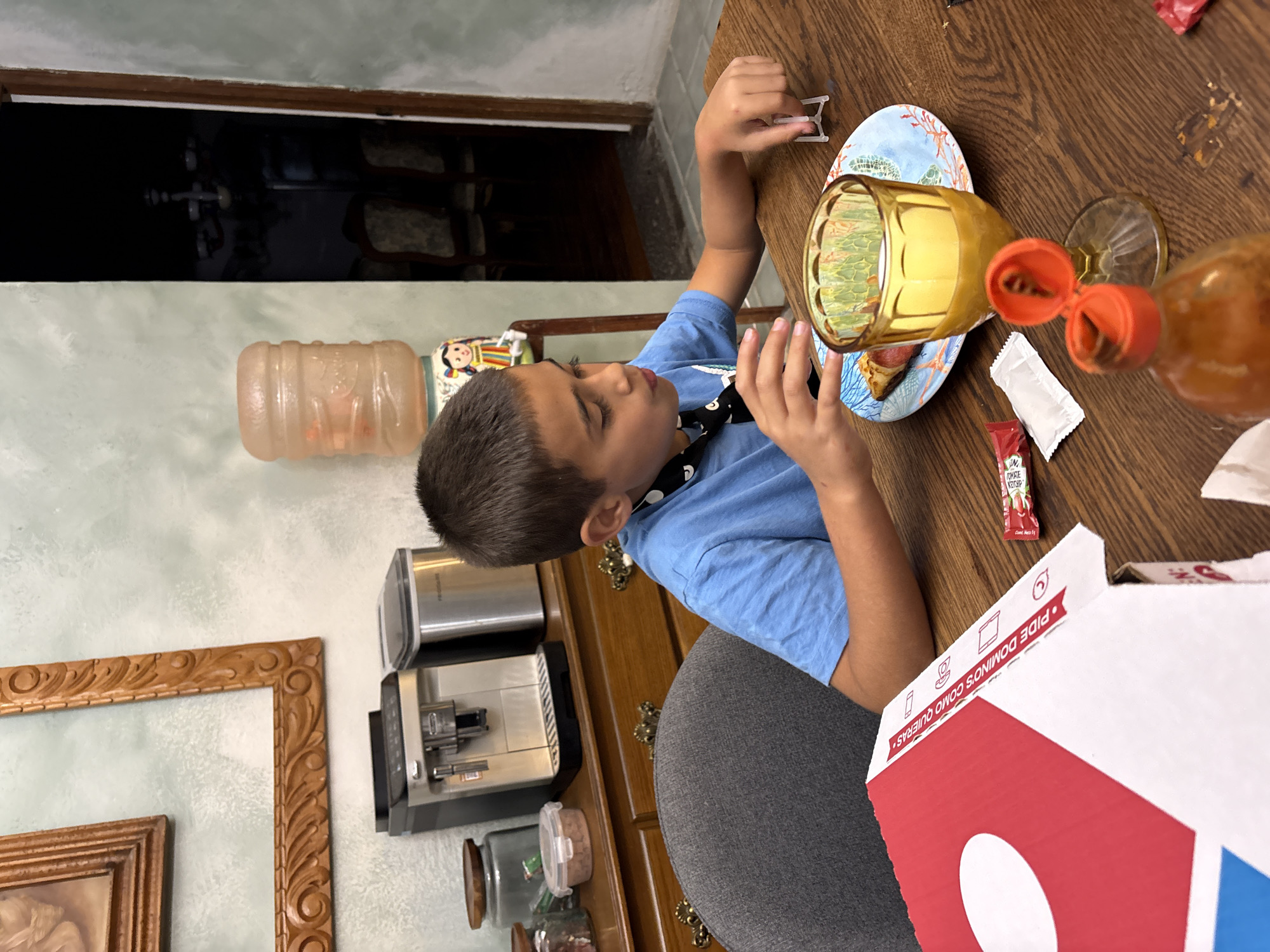 A boy wearing a blue t-shirt and a black and white patterned tie is sitting at a wooden table. On the table are a blue and white plate with what appears to be pizza, a tall glass of yellow liquid, a hot sauce bottle with the cap open, a packet of ketchup, a packet of what might be parmesan cheese, and a Domino's pizza box. The boy is looking down and appears to be engaged with something on the plate. Behind the table is a wooden cabinet with decorative pulls. On top of the cabinet is a water cooler and a coffee machine. A picture frame hangs on the wall above the cabinet. The location is in the city of Puebla, Mexico.