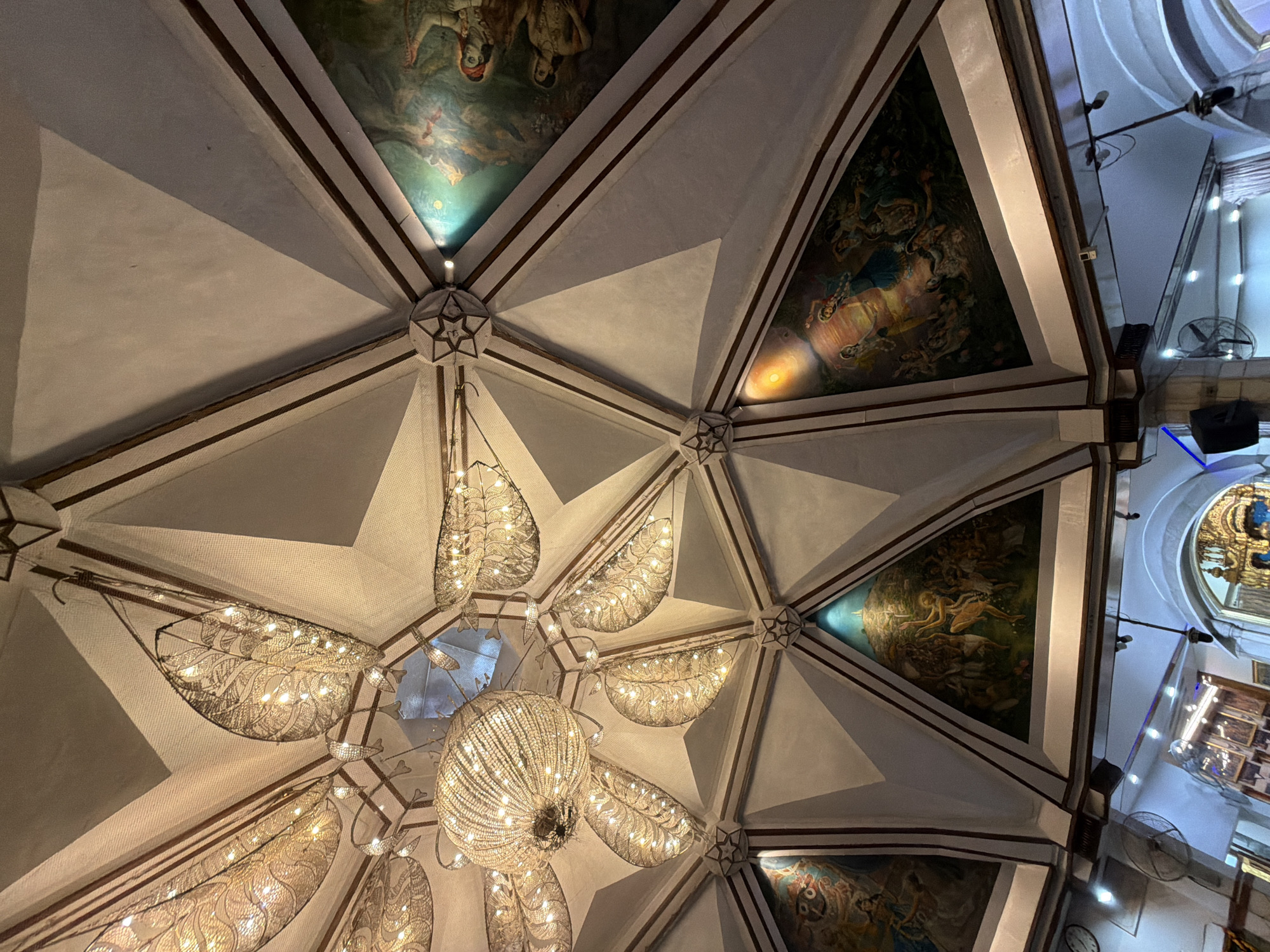 This is a low-angle, indoor shot of a domed ceiling in what appears to be a place of worship or a richly decorated historical building, possibly in Delhi, India. The ceiling is divided into triangular panels by dark brown wooden beams. Each panel features a painting depicting figures, possibly from Hindu mythology or religious narratives, with vibrant colors and detailed scenes.
Suspended from the center of the ceiling is a large, intricate chandelier composed of many small, bright lights. Around the chandelier, and integrated into the ceiling's structure, are several decorative light fixtures resembling large, stylized leaves or feathers, also illuminated with numerous small lights. These fixtures create a warm, ambient glow.
The overall impression is one of grandeur and artistic detail. The lighting suggests it is likely evening or indoors with artificial light being the primary illumination source. The paintings on the ceiling are the most prominent subjects, showcasing artistic skill and cultural significance. The architecture of the ceiling itself, with its geometric divisions and central chandelier, is also a key feature. There are no visible text elements in the frame.