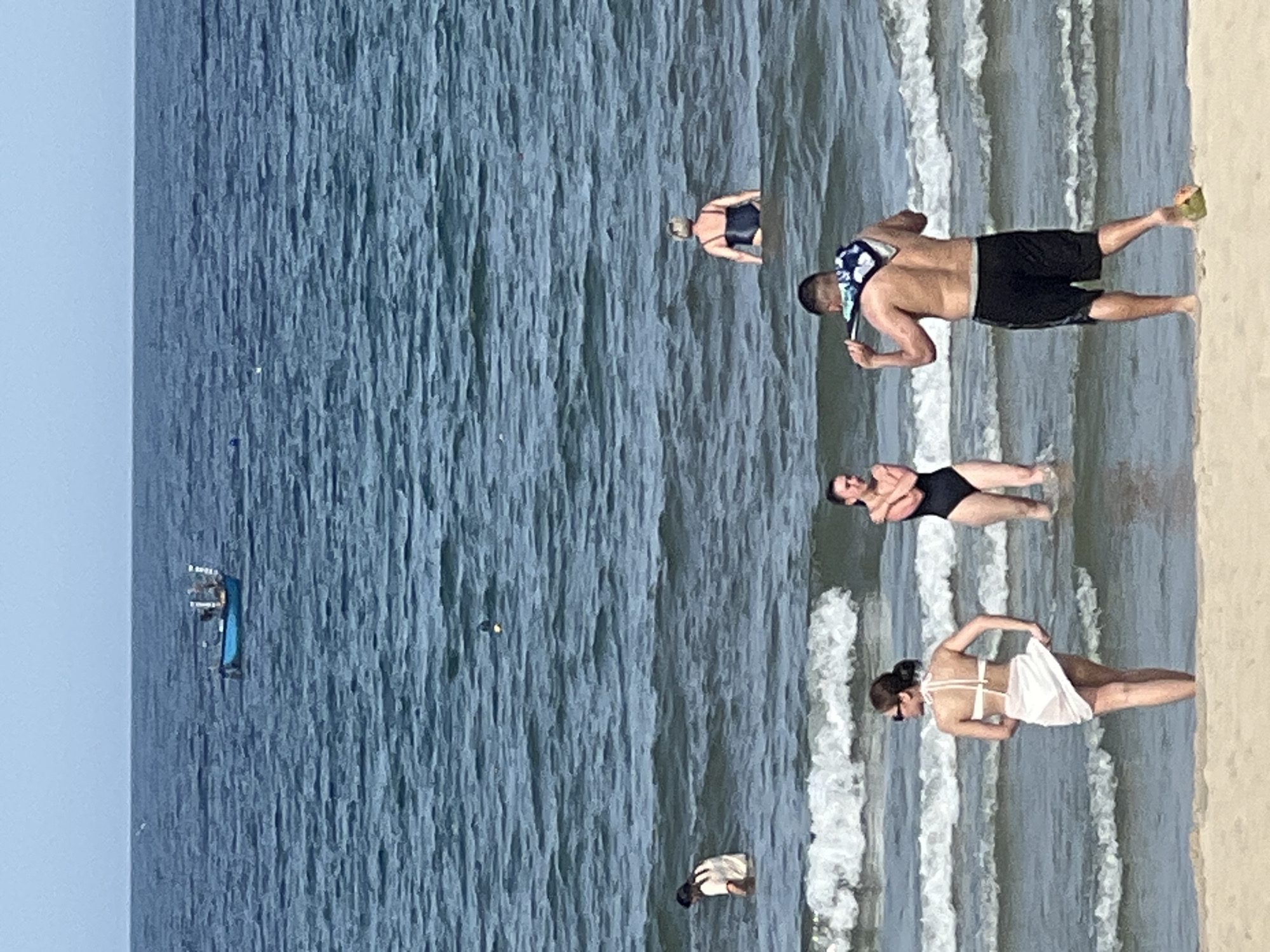 The image shows a view of the ocean from a beach in Đà Nẵng, Vietnam. Several people are in the water. In the foreground, a woman in a white bikini and sarong stands facing away from the camera. To her right, a man in black shorts stands with his back to the camera, a towel draped around his neck. Further back in the water, a woman in a black swimsuit is visible. To the left, another person is partially submerged in the water. In the distance, a small blue boat is visible on the horizon. The water is blue with moderate waves and white foam. The sky is a pale blue. A halved coconut is visible on the sand in the lower right corner of the image.