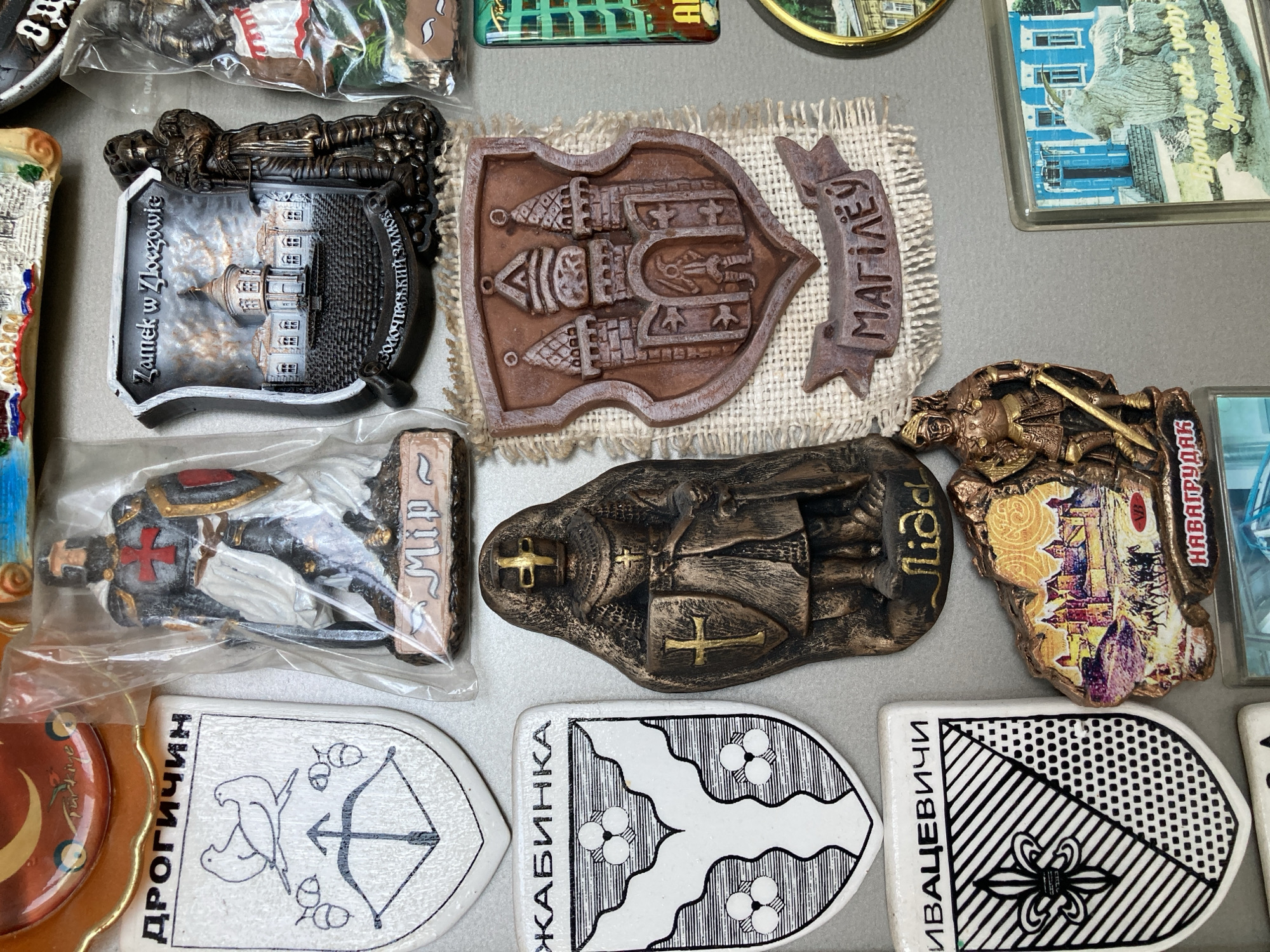 This is a close-up, high-angle shot of a collection of refrigerator magnets, likely displayed on a metallic surface. The magnets are varied in size, shape, and material, featuring different designs and inscriptions. Some magnets depict coat of arms with shields and intricate patterns, while others feature miniature figurines of knights and castles. There are also magnets with text in what appears to be Cyrillic script. The background is a plain, light-colored surface, possibly a refrigerator door. The overall impression is of a display of collectible souvenirs, possibly from various locations.