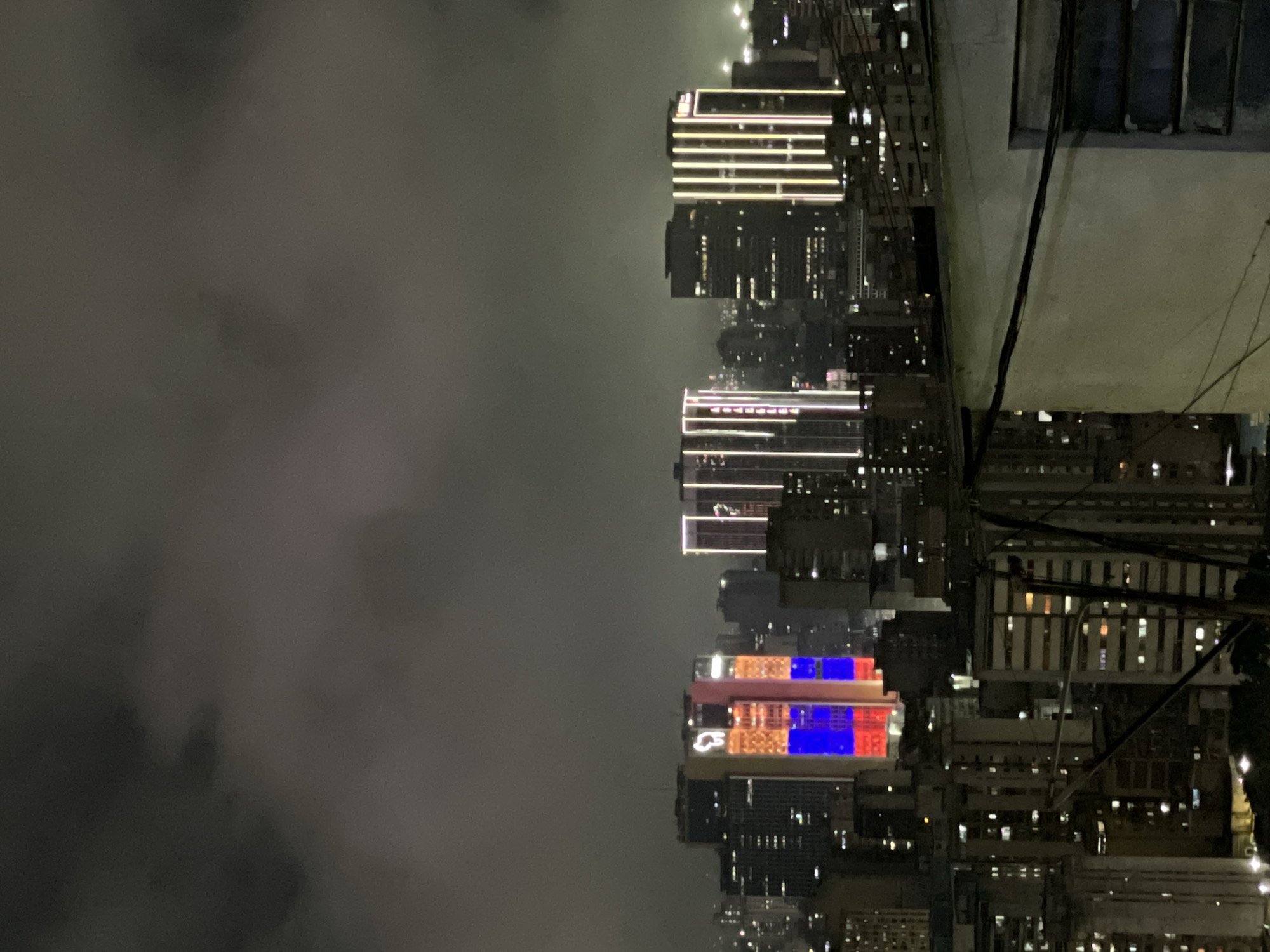 The image captures a night scene in Caracas, Venezuela, featuring a cityscape shrouded in fog. Tall buildings with illuminated windows and vibrant displays of red and blue lights dominate the view. A dense network of power lines extends across the foreground. A residential building with a window is positioned in the upper right corner. The overall atmosphere is hazy, with the city lights blurring into the fog.
