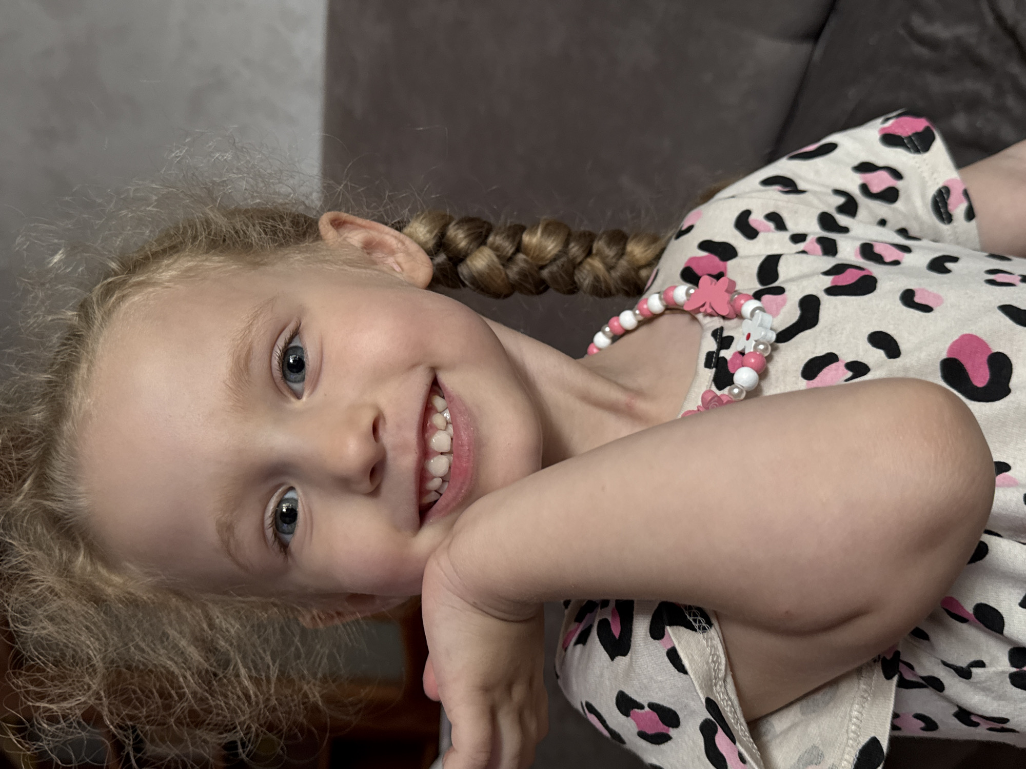 The image shows a close-up, rotated portrait of a young girl with blonde hair and blue eyes, smiling. She is wearing a white top with a pink and black leopard print pattern and a beaded necklace with pink and white beads and a flower charm. Her hair is styled in a braid, with some looser curls framing her face. The background is a blurred, neutral color, suggesting an indoor setting, possibly a living room or sofa. The lighting appears to be natural or soft indoor lighting, and the time of day is not discernible. The girl's expression is happy and playful. There is no visible text in the image. The location context provided is Chaikovsky, Russia.
