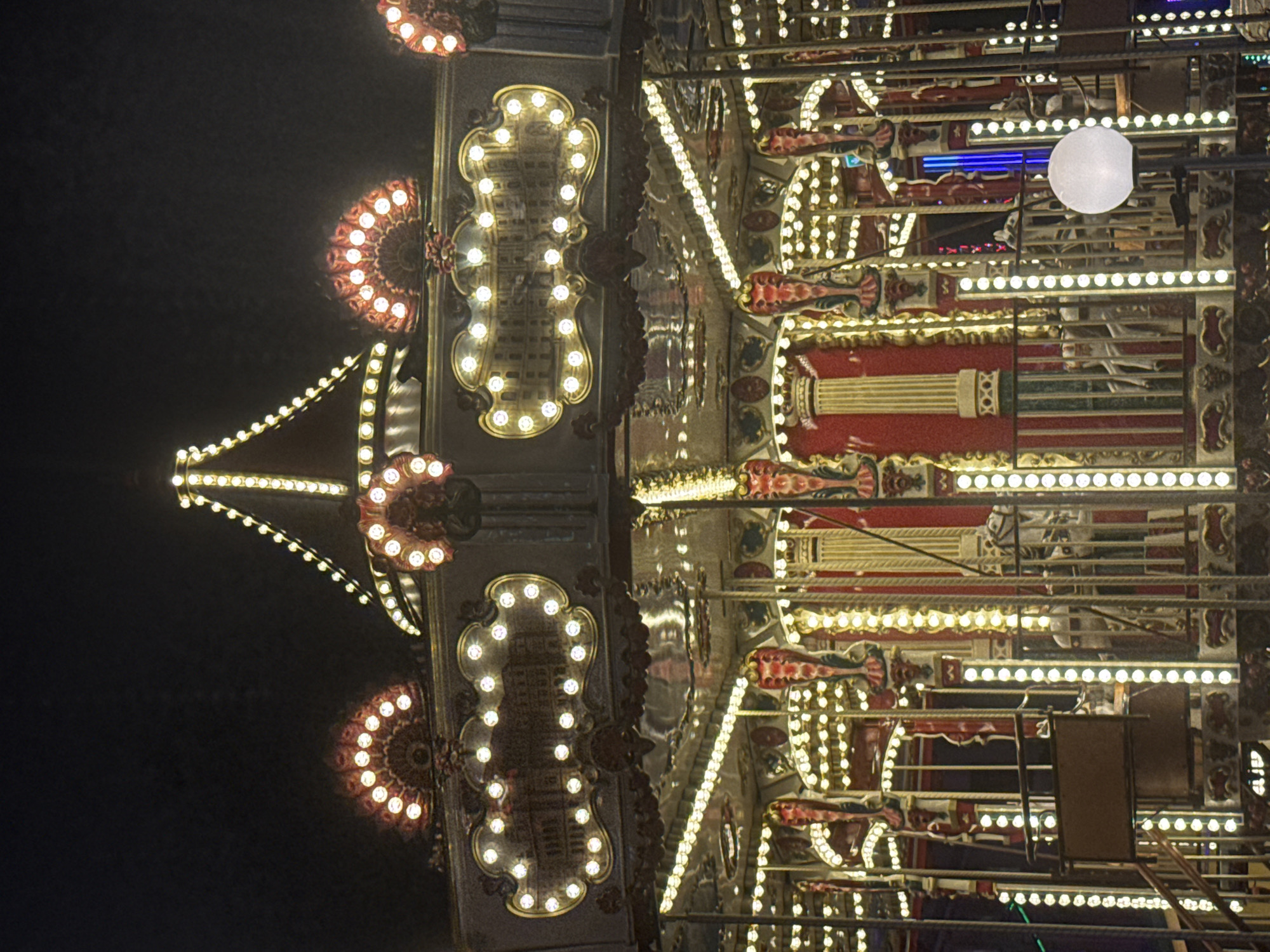 This is a photograph of a brightly lit carousel at night. The carousel has a circular design with multiple tiers, all adorned with numerous yellow and white lights. The upper section features decorative fan-shaped light displays and what appears to be a mural of a building within an ornate frame. The lower section of the carousel reveals the structure of the ride, with decorative columns, railings, and what seem to be the backs of carousel horses. The overall impression is one of a festive amusement ride illuminated against a dark, nighttime sky. The image does not provide any explicit visual cues or landmarks to definitively place it in Vienna, Austria.