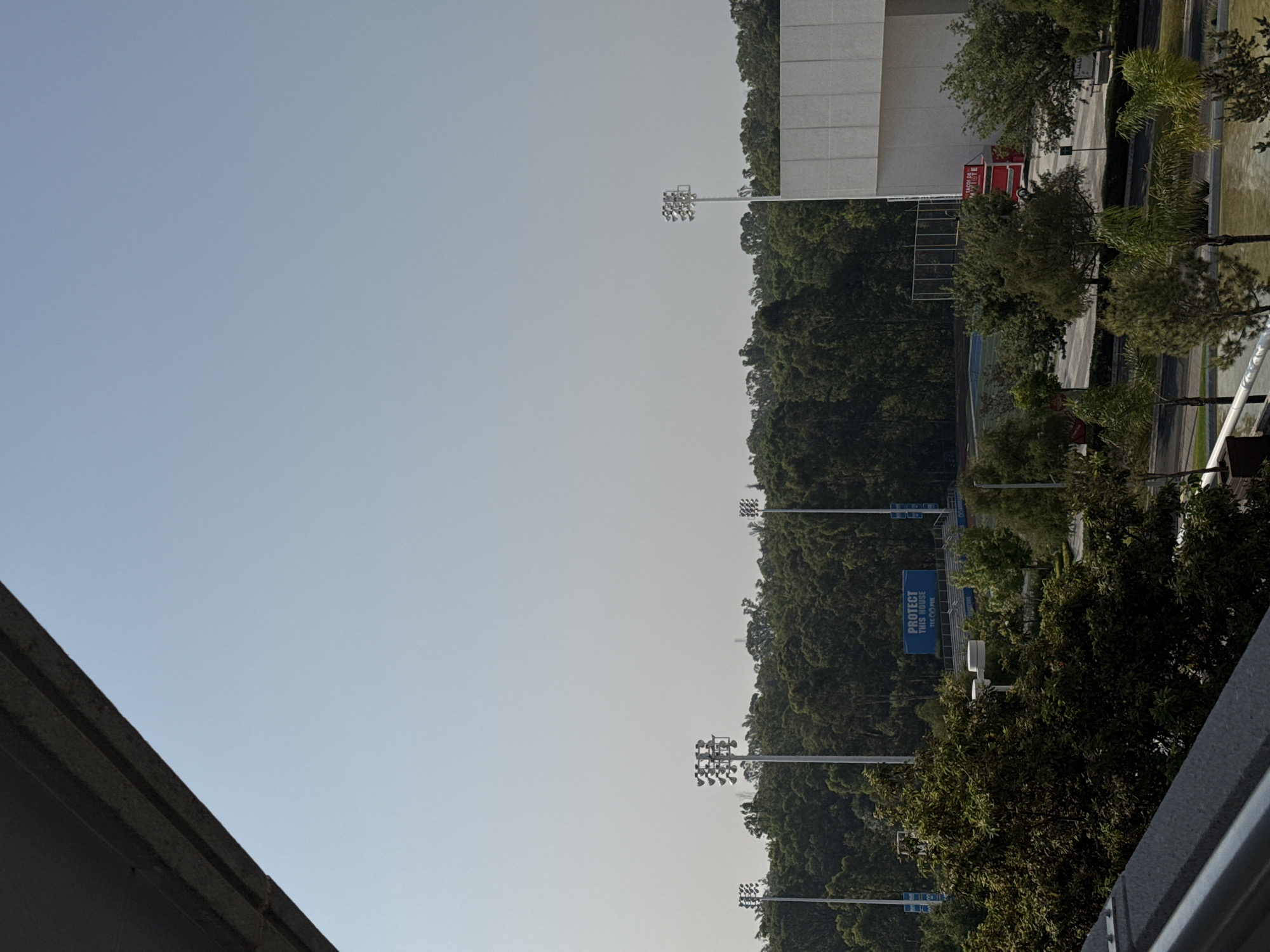 The image shows a view in Puebla, Mexico, likely taken from an elevated position. The scene includes a sports stadium with lighting towers, partially obscured by dense trees. A large white building is visible in the background. The stadium has blue signage. The sky is pale and clear.