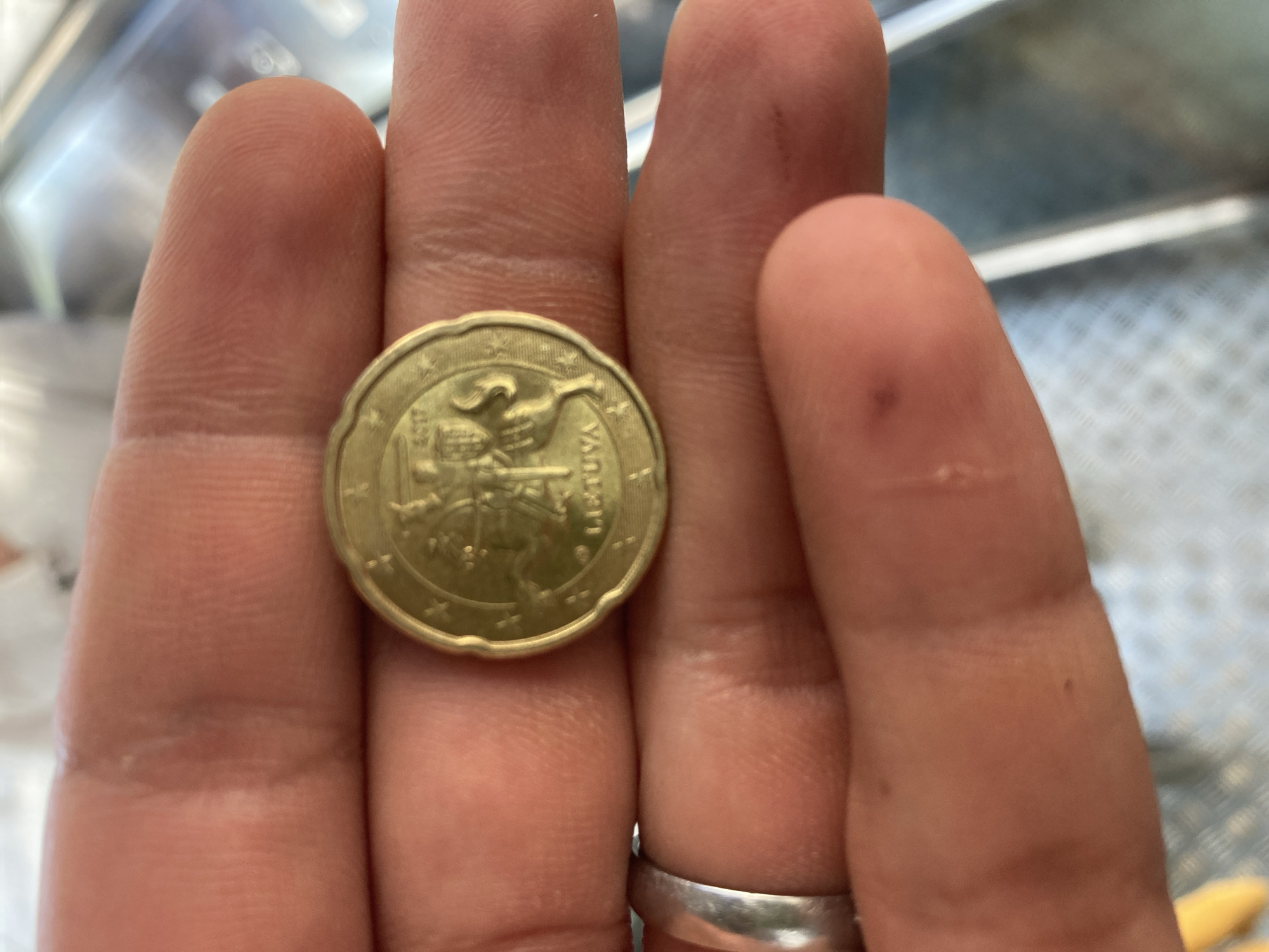 A close-up image captures a person's hand holding a gold-colored coin. The hand is positioned with fingers spread, and a silver-toned ring is worn on the ring finger. The skin of the hand shows natural creases and a few small, dark marks.
The focal point is the coin, which displays the Vytis, the national emblem of Lithuania, featuring a mounted knight on horseback. The word "LIETUVA" is clearly visible along the right edge of the coin's face, confirming its origin. The year "2017" is partially visible near the bottom left of the coin's design. A ring of stars, characteristic of Euro coins, encircles the Vytis emblem.
The background is softly blurred, showing indistinct light and dark areas, possibly indicating an indoor setting with some metallic or patterned surfaces. The lighting appears bright and even, illuminating the hand and coin distinctly. No specific activity beyond holding the coin is depicted. The time of day and weather are not discernible.