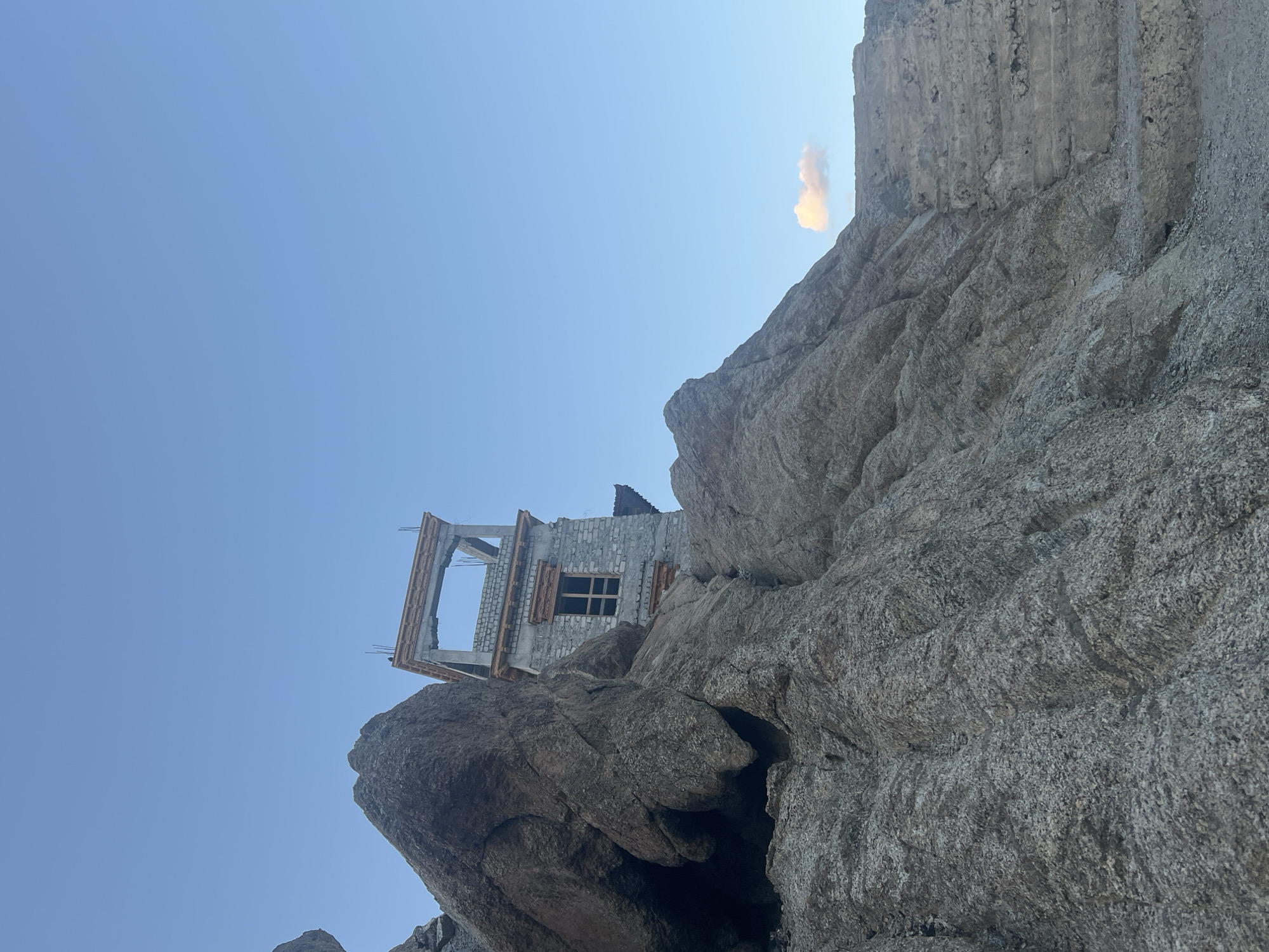 The image shows a view of the Leh Palace under construction, nestled within the rugged, rocky terrain of Leh, India. The foreground is dominated by large, textured rock formations, casting shadows and creating a sense of scale. Above and behind these rocks, a partially constructed stone building, identified as a part of Leh Palace, is visible against a clear blue sky. The building features traditional architectural elements, including wooden window frames and decorative stonework. A single, small cloud is present in the upper right portion of the sky. The overall scene suggests a remote, high-altitude location with ongoing construction in a historically significant site.