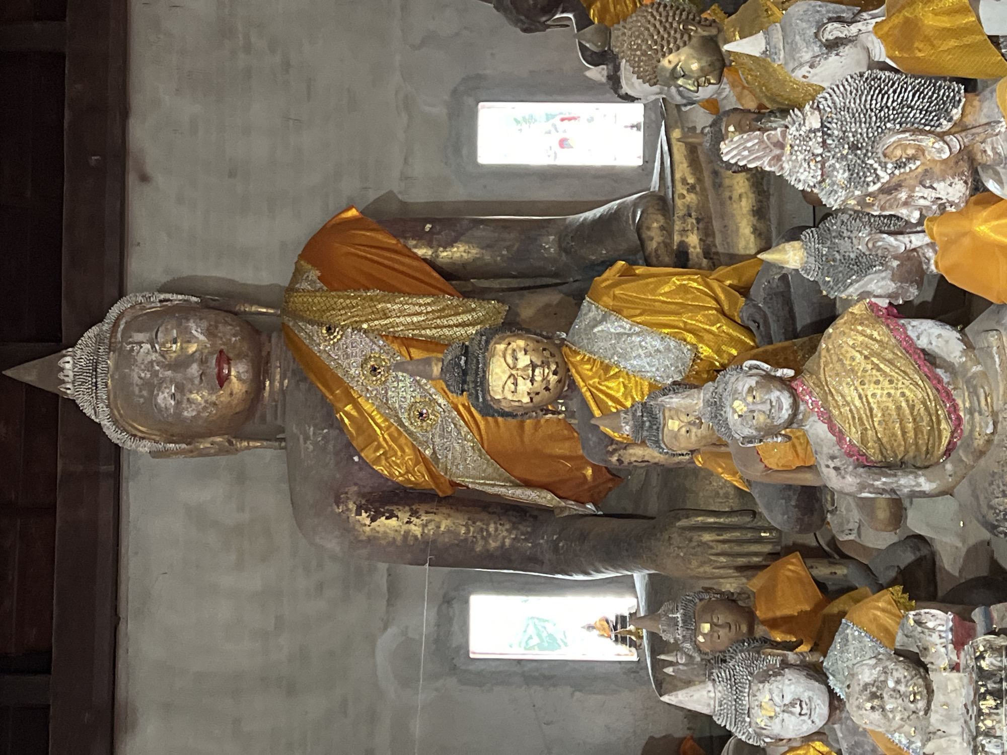The image depicts a collection of Buddha statues within an indoor structure, possibly a temple or shrine, located in the Ko Samui District of Thailand. A large, central Buddha statue, adorned with golden robes, dominates the scene. Smaller Buddha statues surround the central figure, also wearing golden or orange robes and some with silver-colored headdresses. The statues appear aged, with some showing signs of wear and patina. Two rectangular windows with light filtering through are visible in the background, casting a soft glow on the surroundings. The architecture suggests a traditional Thai design, with wooden beams visible at the top of the frame. The overall impression is one of reverence and historical significance.
