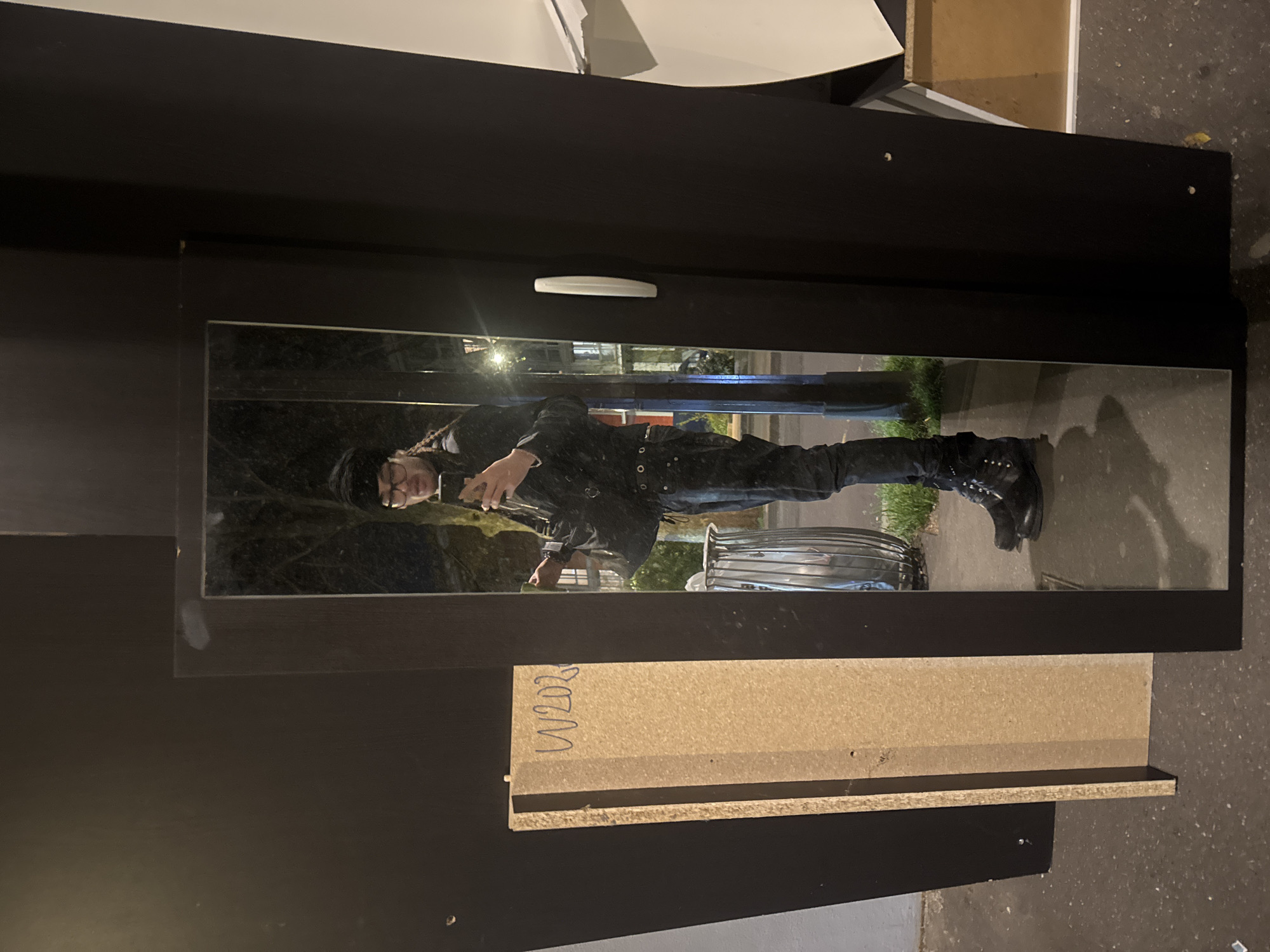 The image shows a person reflected in a mirrored wardrobe door. The person is wearing a black hat, glasses, black jacket, and black boots. The reflection reveals a city street scene with trees, buildings, a streetlight, a trash can, and a sidewalk. The surroundings indicate an urban environment, potentially Paris, France. The wardrobe appears to be in a state of partial disassembly, with a drawer and other wooden pieces visible.