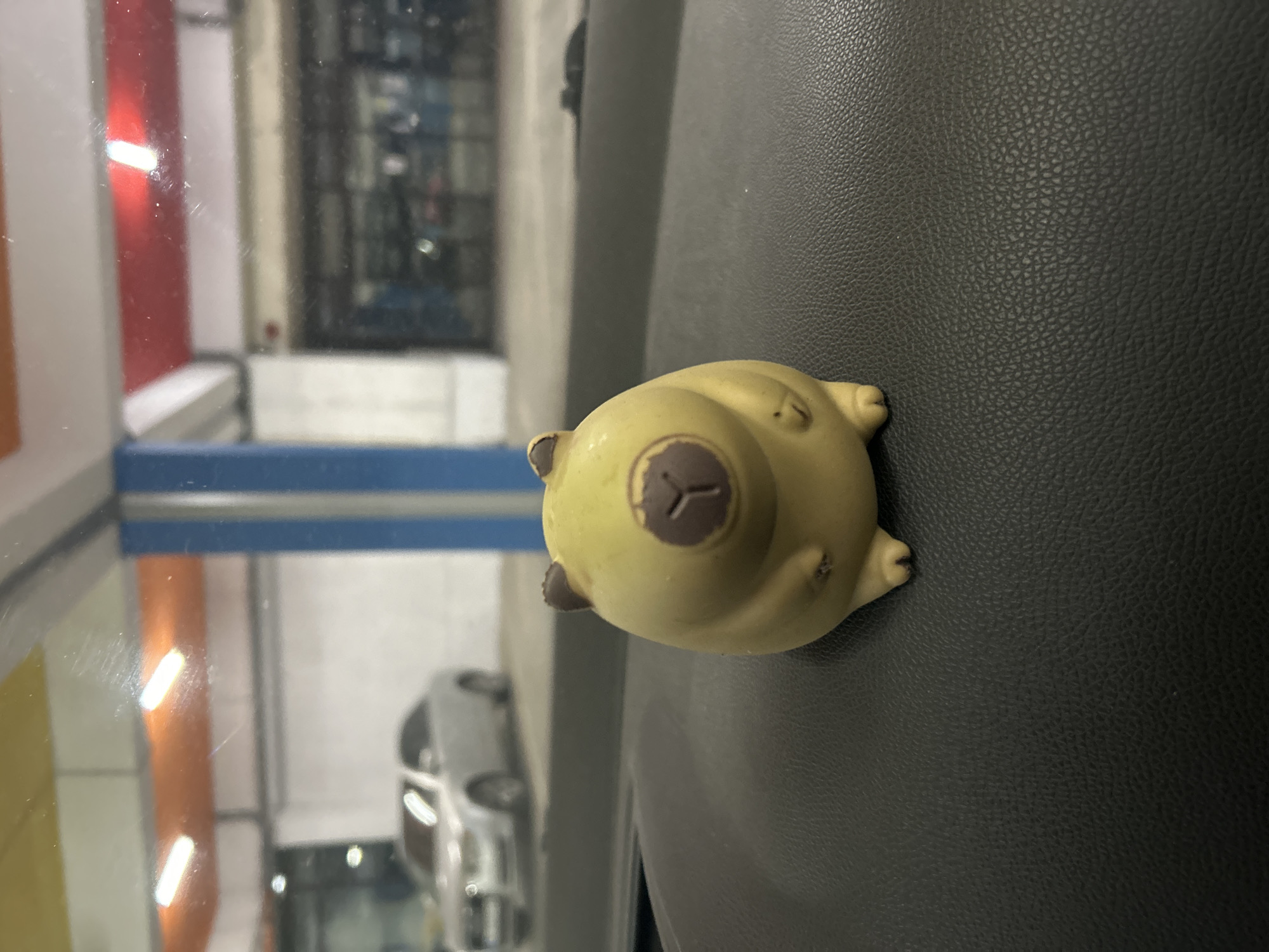A yellow, stylized bear figurine with a brown nose is positioned on a black, textured surface. The background shows an out-of-focus reflection of a street scene with a multi-story building, a blue structure, and what appears to be a car. The location is likely Varna, Bulgaria. No people are visible.