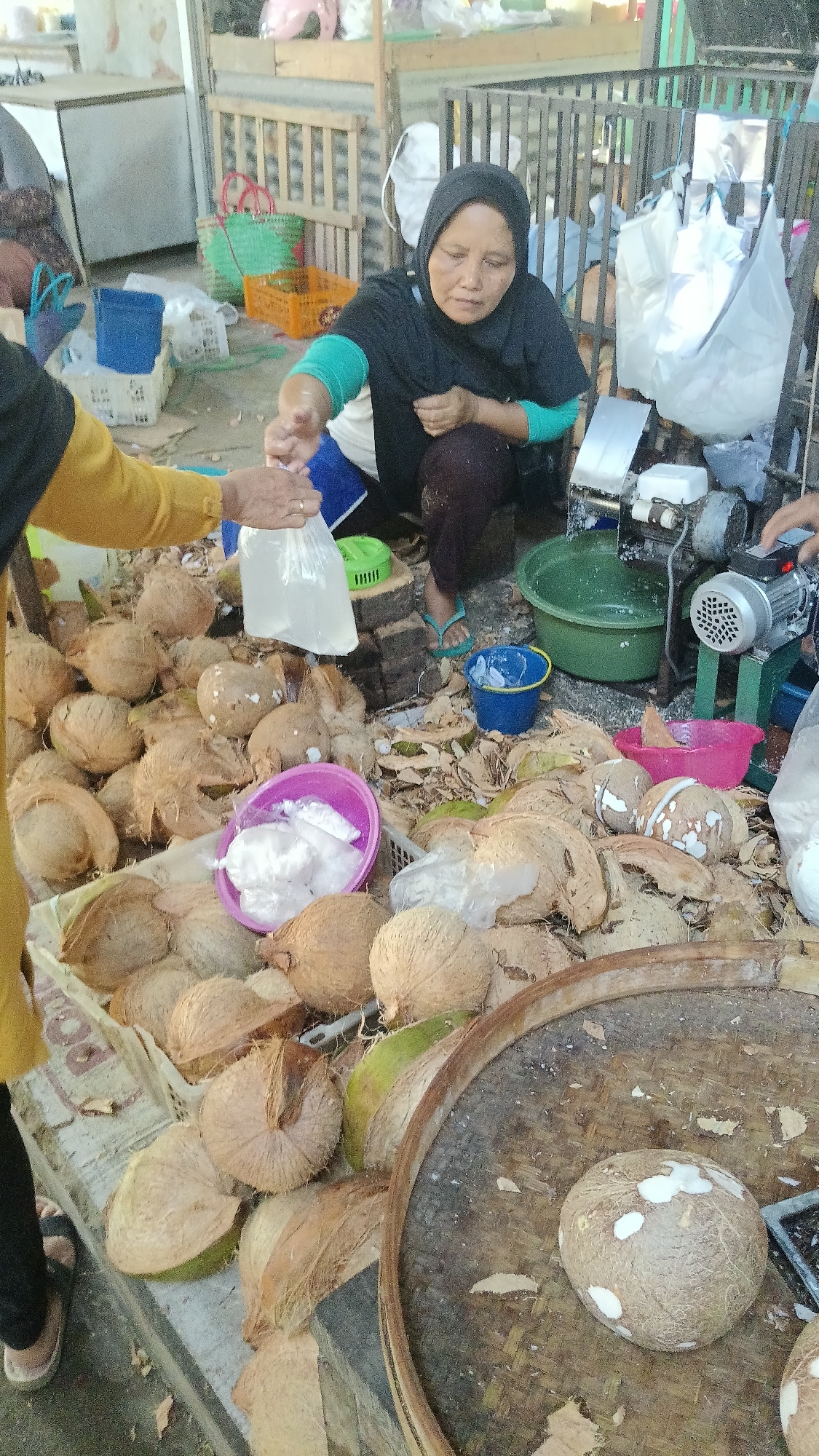 A market scene in Dukuh, Indonesia, shows a woman wearing a black hijab and a black shirt with teal sleeves, crouched down and in the process of preparing coconuts. She is holding a clear plastic bag filled with coconut water, apparently having just collected it. In front of her are many whole coconuts and coconut husks, indicating she is processing them. A purple bowl with shredded coconut is visible, as are several other bowls and buckets in various colors (green, blue, pink). A coconut-shredding machine is present to the right, along with a large green basin. Another person's hand is visible, also interacting with the coconuts. The ground is covered with coconut debris. The overall impression is of a busy, open-air market stall specializing in coconut products.