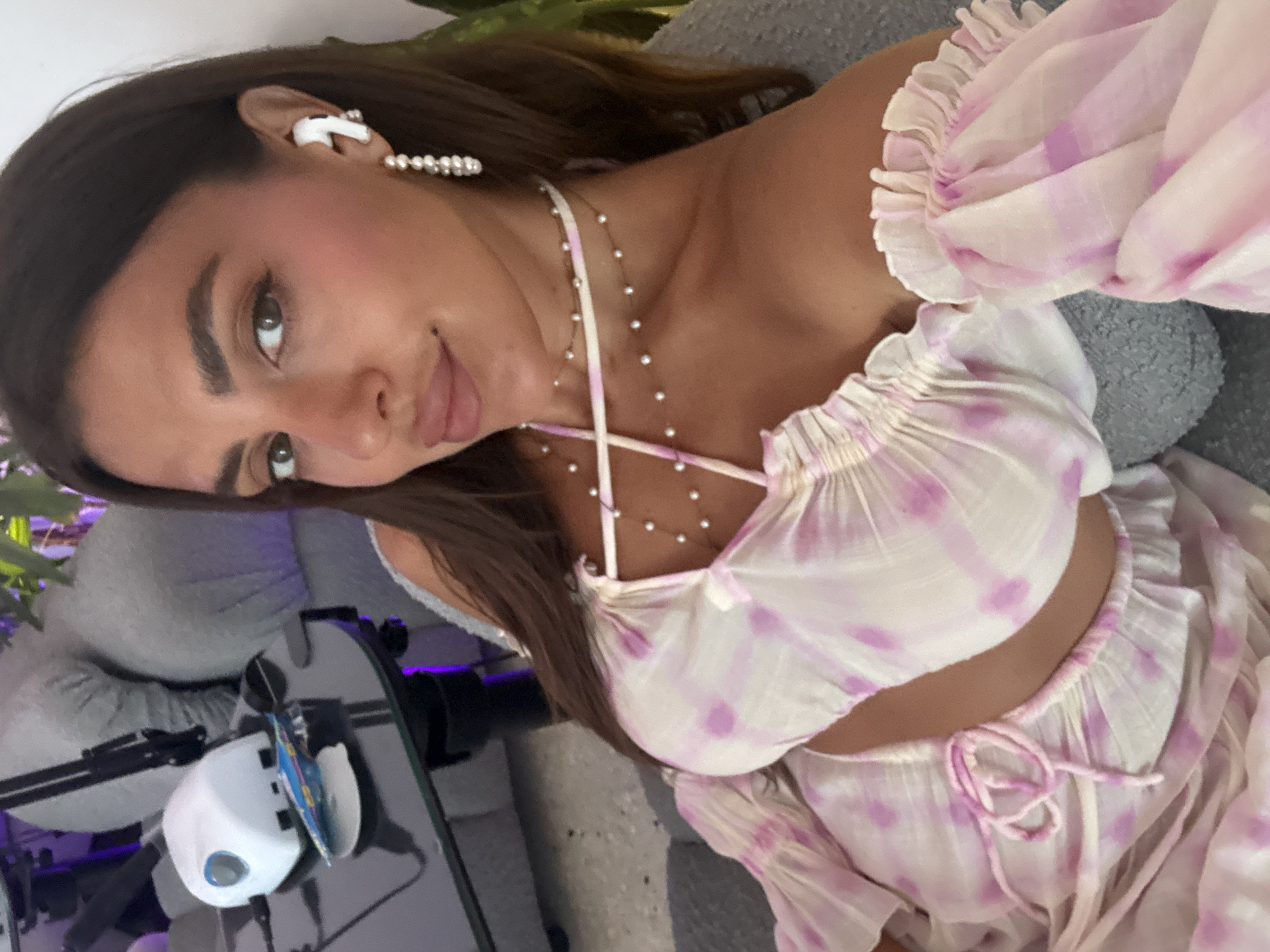 A young woman is depicted in a close-up, angled selfie, appearing to be indoors in North Kuta, Indonesia. She has long, dark brown hair and dark eyes, looking directly at the camera with a relaxed expression and a subtle, coy smile. She is dressed in a two-piece set featuring a white fabric with a soft pink or purple tie-dye pattern. The top is an off-the-shoulder crop top with ruffled elasticated sleeves and a ruffled hem, revealing her midriff. A matching lower garment, possibly shorts or a skirt, with a drawstring tie, is also visible.
Her accessories include a white wireless earbud placed in her right ear, complemented by a delicate pearl ear cuff or drop earring hanging just below it. Around her neck, she wears a fine, multi-strand necklace adorned with small white, pearl-like beads.
The setting appears to be an indoor space, possibly a home or hotel room. She is seated against a grey upholstered surface, which could be a sofa or headboard. To her left, on a dark table, rests a white and grey electronic device, possibly a small fan or diffuser, with a blue button and a small card inserted into it. Hints of green foliage from a plant are visible in the background, along with subtle purple ambient lighting, creating a relaxed atmosphere. There is no visible text in the image.