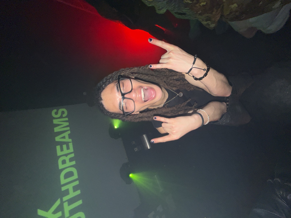A person with dreadlocks and glasses poses, making a "rock on" hand gesture with both hands. They wear dark nail polish, a necklace, and several bracelets. The background is dark with green and red lighting. A screen displays the word "YOUTHDREAMS" in green. People can be seen in the blurred edges.