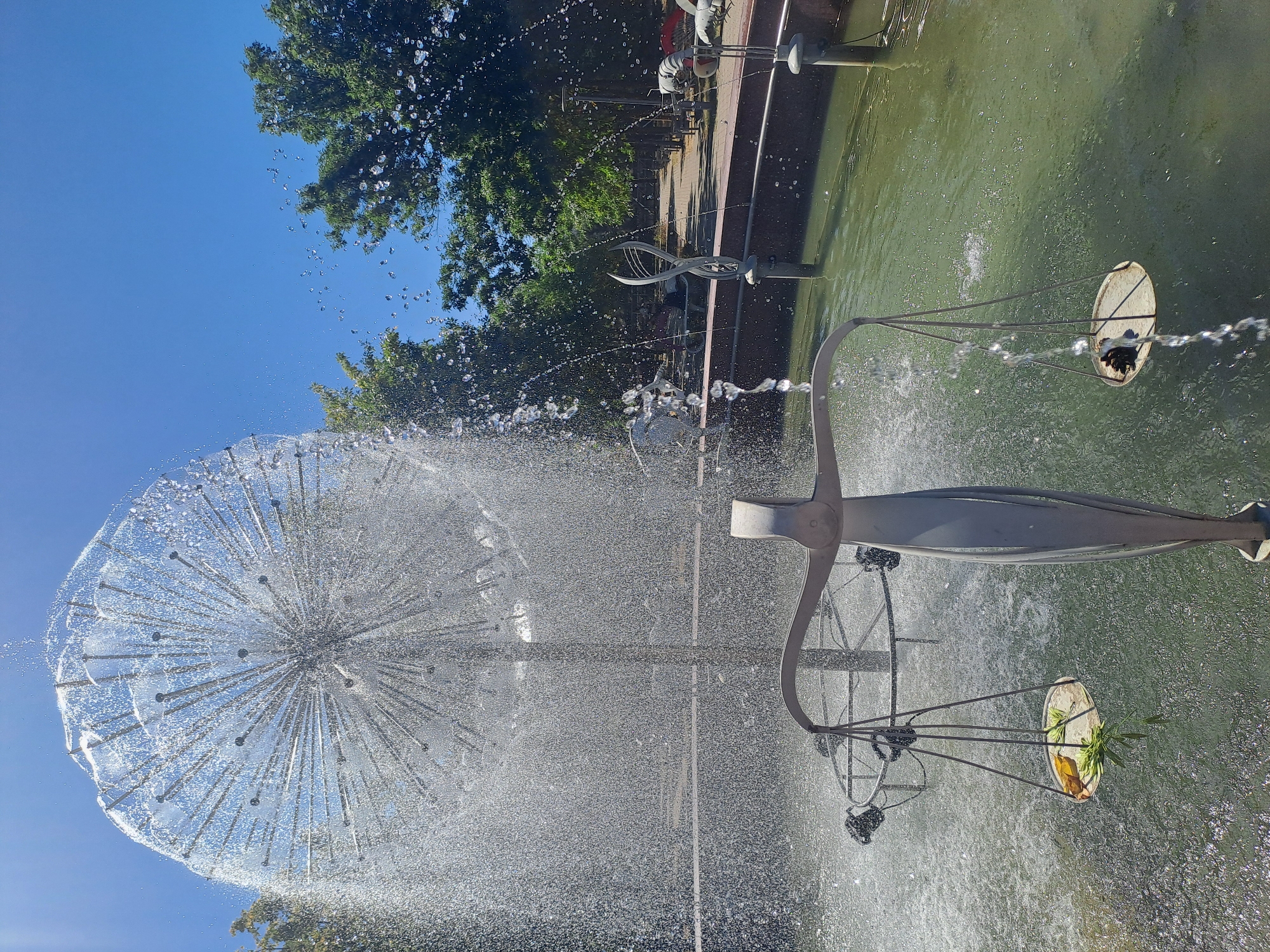 This image depicts a public fountain with a unique sculptural element resembling a dandelion seed head, from which water sprays outwards, creating a shimmering effect against a clear blue sky. In the foreground, a stylized scale of justice sculpture stands partially submerged in the fountain's green water. One pan of the scale holds a few pieces of what appear to be green leaves and a yellow object, while the other pan holds a dark, pinecone-like object. The fountain is situated in an outdoor area with trees and a paved walkway visible in the background, suggesting a park or public square. The overall impression is one of a sunny day in an urban recreational space.
The specific location in Chișinău, Moldova, is the "Fântâna cu Scara" (Fountain with Scales) located in the Valea Morilor Park. This fountain is a notable landmark in the park, often featuring the scale of justice sculpture, though its contents can vary. The park itself is a popular recreational area in the city, known for its lake and green spaces.