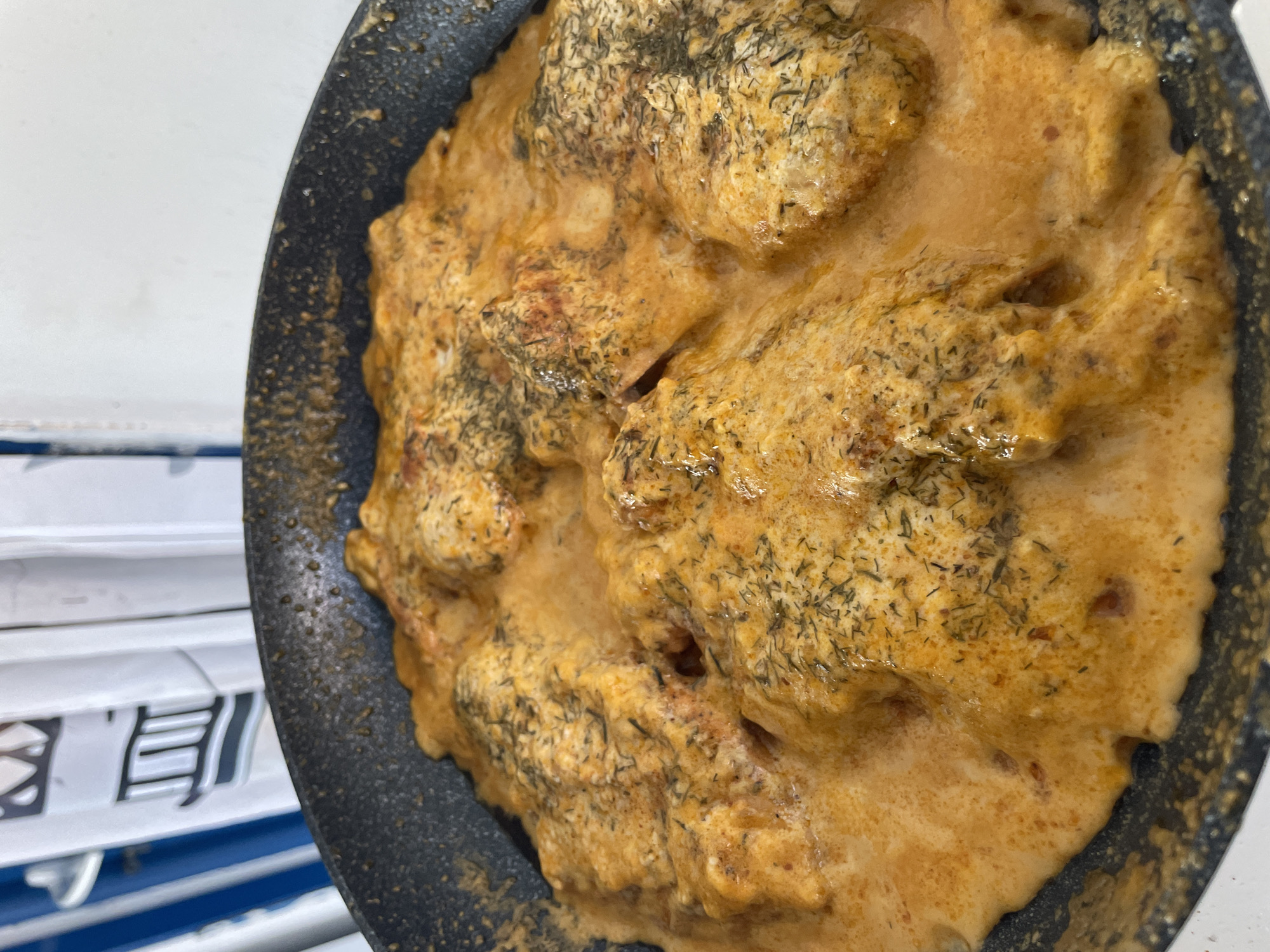 This is a close-up, overhead shot of food being cooked in a dark skillet. The food appears to be a creamy, orange-hued dish with visible pieces of what might be chicken or meatballs, coated in a rich sauce and sprinkled with dried herbs, likely dill. The sauce is thick and glossy, with some areas showing a slightly bubbly texture, suggesting it's actively simmering.
The skillet itself is black and has a textured surface. Part of the background shows a white surface with some horizontal blue stripes and what appears to be text in a foreign language, possibly in Asian script, printed on white rectangular elements. This suggests the cooking might be taking place indoors, perhaps in a kitchen or an establishment where such signage is present.
There are no people or discernible faces visible in the image. The lighting is bright and even, casting no strong shadows, which could indicate artificial lighting or an overcast day. The focus is entirely on the food, highlighting its texture and appetizing appearance. The presence of the foreign text in the background might hint at the location being a restaurant, or a place where international products are sold.
Given the provided location context of Skala Avdiron, Greece, the foreign text is unusual, unless it refers to imported ingredients or a specific type of cuisine being served. The image itself doesn't provide any specific clues about the time of day or weather conditions. The overall impression is a visual of a savory dish being prepared.