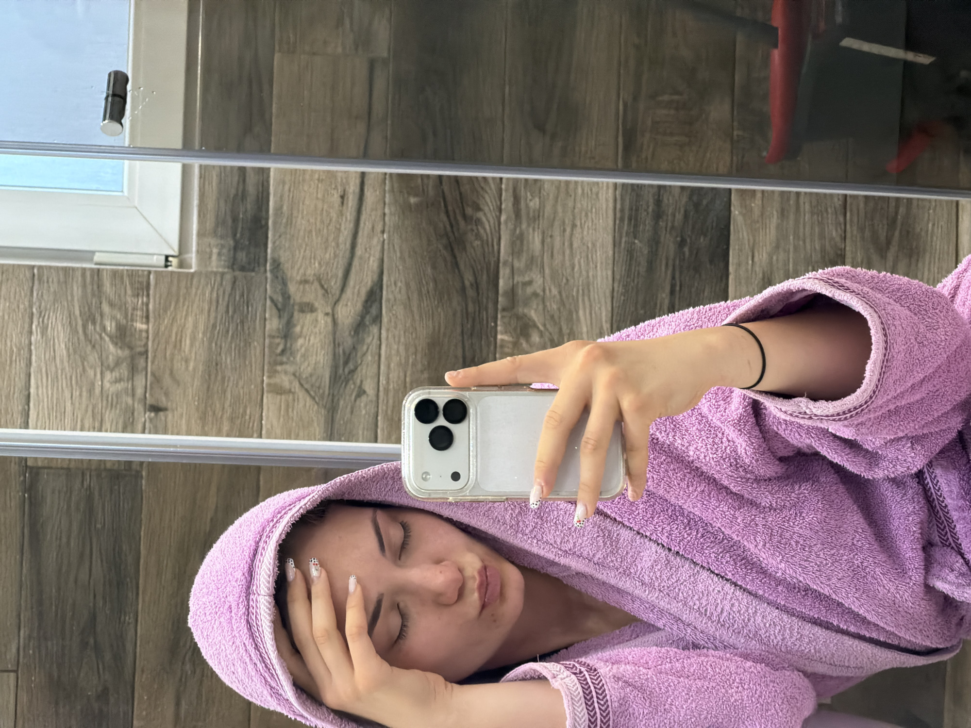 A woman is lying down in a bathroom in Buhovo, Bulgaria. She is wearing a purple hooded bathrobe and holding a white smartphone up to take a selfie. Her eyes are closed and she is pursing her lips. Her nails are painted with a white base and small black spots. She has a black bracelet on her wrist. The bathroom has wood-paneled walls and a glass shower enclosure. A red object is faintly visible in the reflection of the shower glass.
