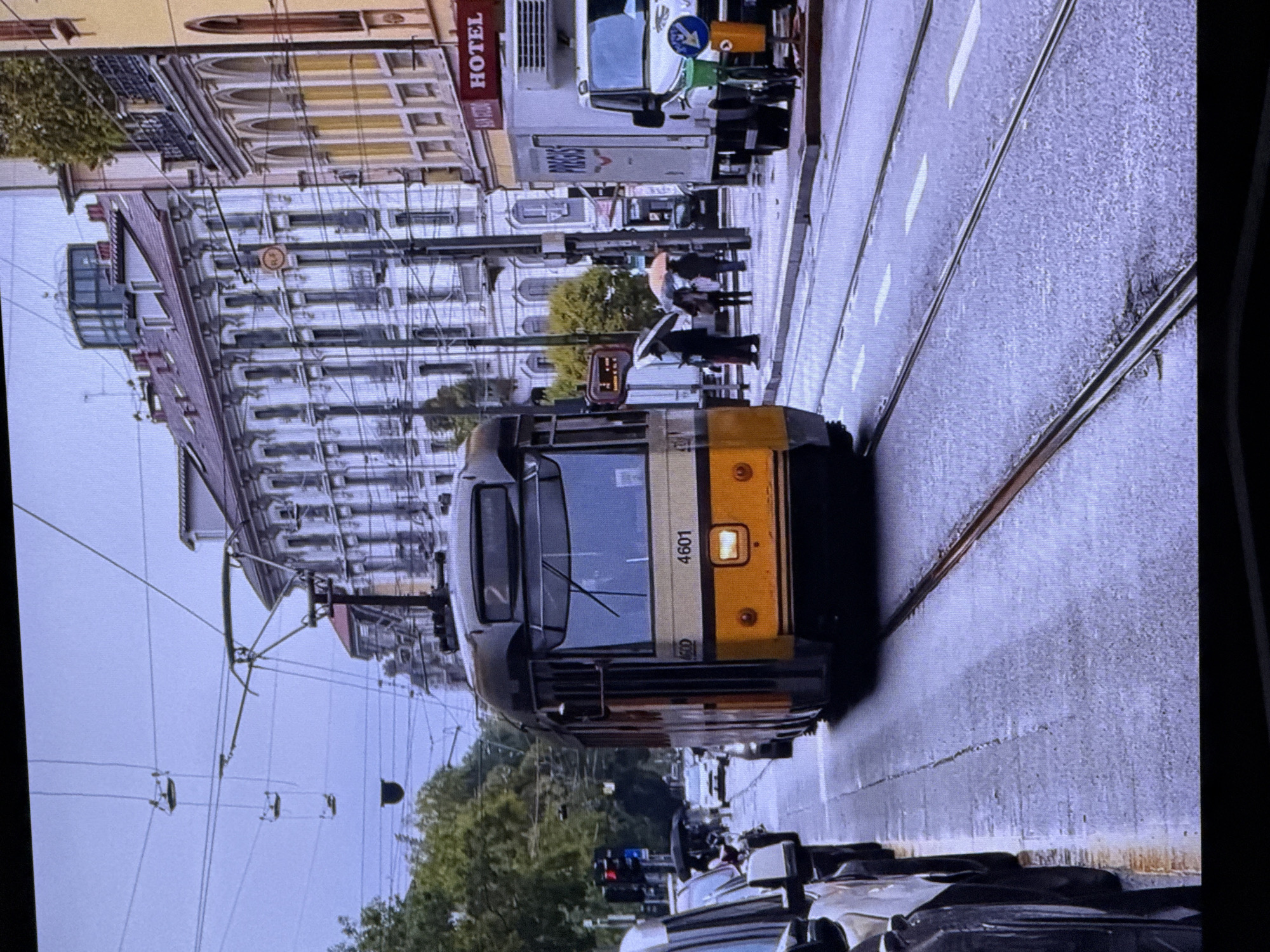 This image depicts a tram on a wet city street, with several people holding umbrellas. The tram, numbered 4601, is yellow and black and appears to be in motion. The street is lined with buildings, some of which have balconies and elaborate architectural details. There are also overhead wires for the tram's power supply, and several cars are parked along the side of the road. A white refrigerated truck is parked on the right side of the street. The sky is overcast, suggesting cloudy or rainy weather. The scene appears to be set in an urban environment with a European architectural style. There is no indication that this scene is located in Dubai, United Arab Emirates.