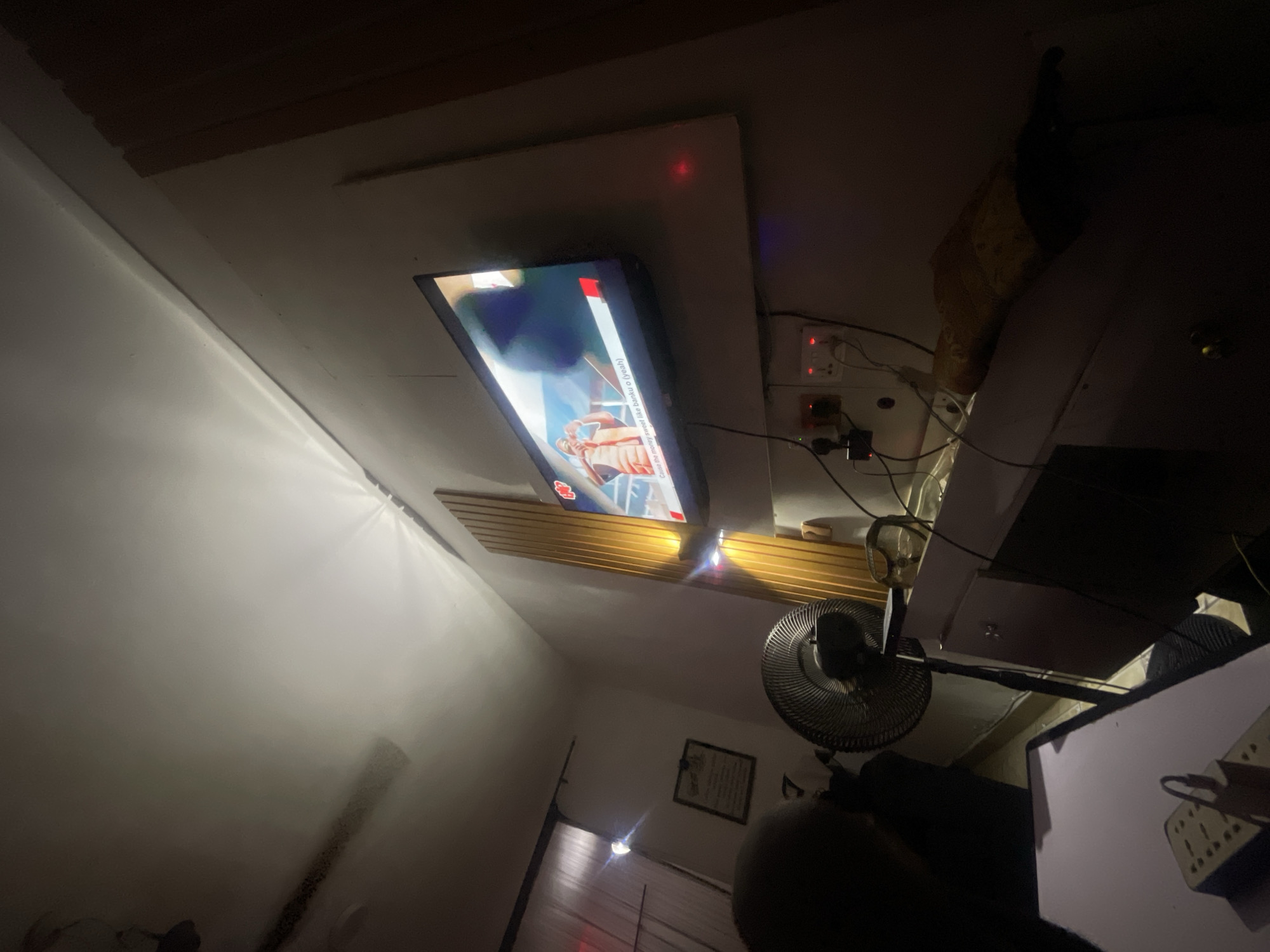 The image shows an interior space in Lokoja, Nigeria. A television is mounted on the ceiling, displaying content. Adjacent to the television is a series of electrical outlets and a surge protector. A fan is positioned in the foreground, partially obstructing the view. A desk is partially visible on the right. The lighting in the room is dim. A framed certificate or picture hangs on the wall near the fan.