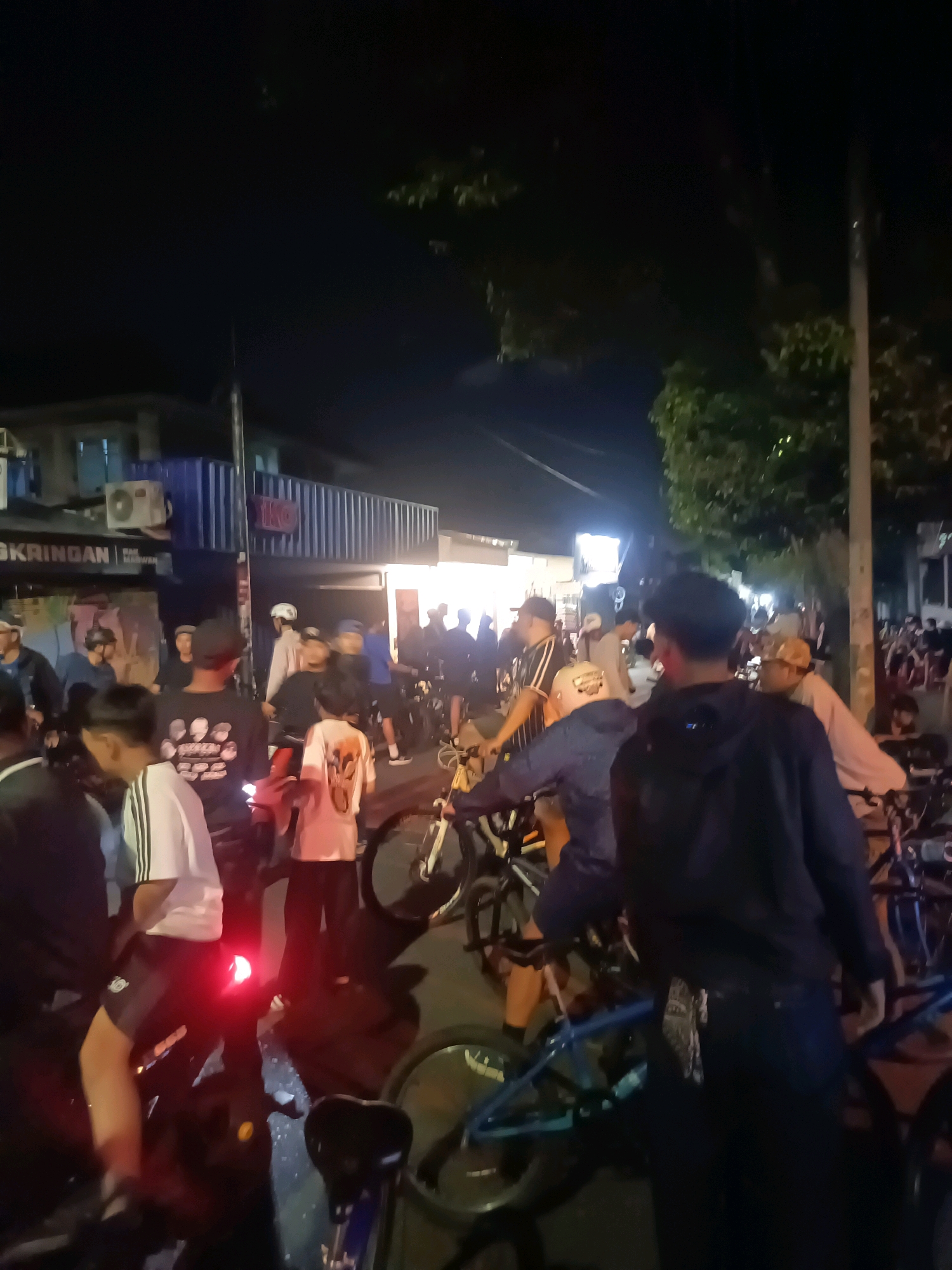 A group of people are gathered on a street at night in Yogyakarta, Indonesia, with many of them on bicycles. The scene appears to be outside a row of buildings that have signs and lights. The individuals are dressed in casual clothing, and some are wearing helmets. Several bicycles are visible, including what appear to be BMX bikes. The lighting suggests it is dark outside, with artificial lights illuminating the area. The overall impression is of a social gathering or event involving cyclists.