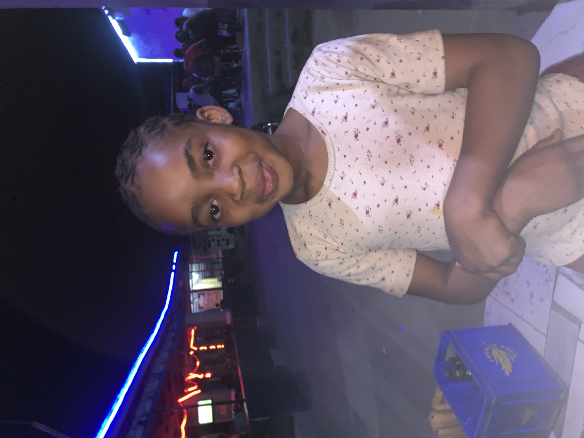 A young person is positioned at the right of the frame, facing the camera with a slight smile. They are wearing a light-colored shirt patterned with small red and green designs. Their arms are crossed in front of them. A blue plastic crate with yellow markings is in the foreground. In the background, there are people gathered and building with neon lights. The location is New Stadium, Nigeria.