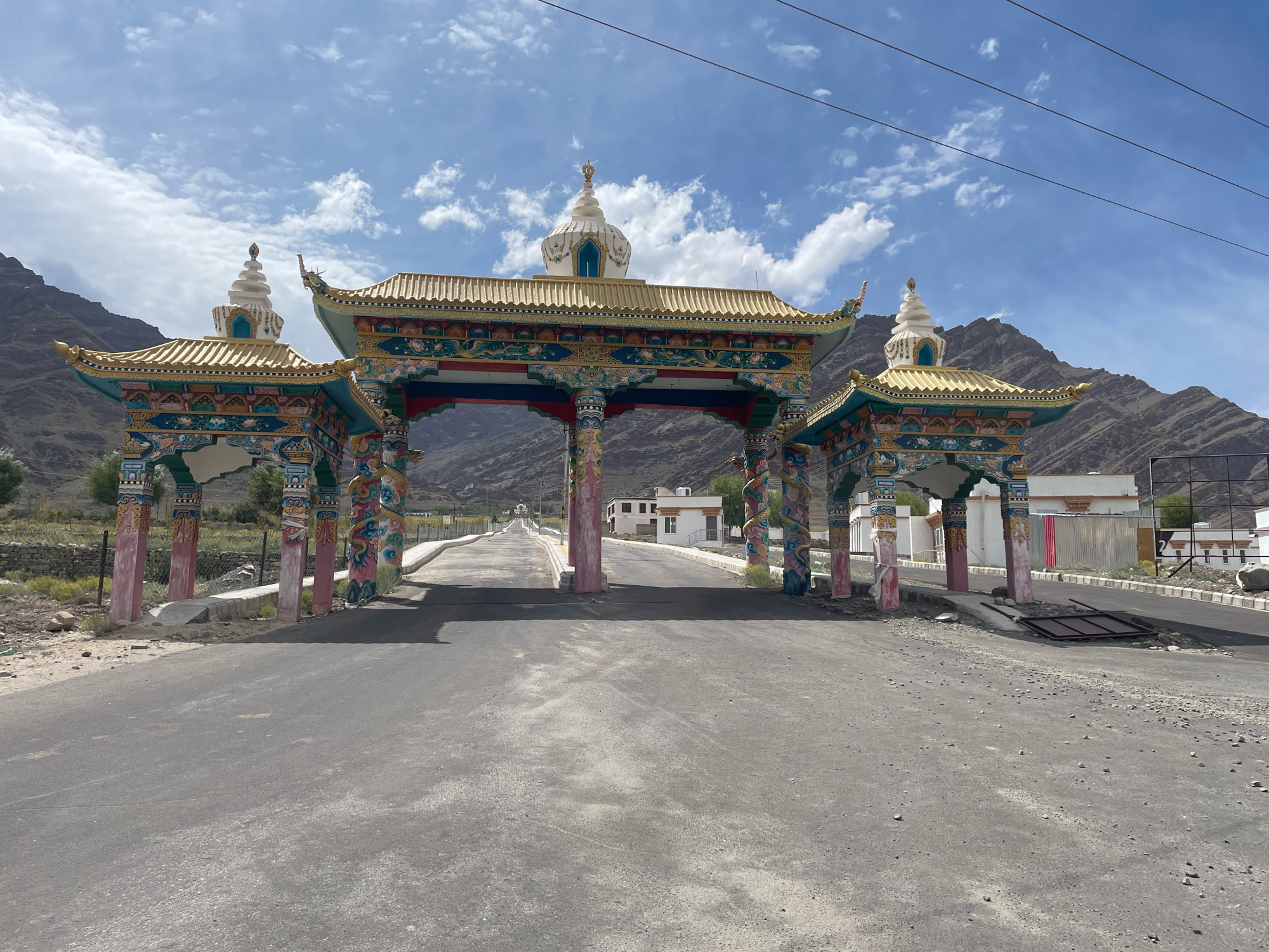 The image shows a wide, paved road leading through a brightly colored, ornate gateway. The gateway is supported by multiple pillars adorned with intricate carvings and paintings of mythical creatures and decorative patterns. The roof of the gateway is golden and features multiple tiers and decorative finials, with stupa-like structures at the highest points. The architecture suggests a Buddhist influence, consistent with the location in Martselang, India, a region known for its monasteries and Tibetan Buddhist culture.
The road itself is asphalt, appearing well-maintained but showing signs of wear, with some patches of gravel and dust along the edges. The gateway is set against a backdrop of barren, mountainous terrain, characteristic of high-altitude regions like Ladakh. The sky above is a vibrant blue, dotted with white, fluffy clouds.
In the background, beyond the gateway, several white buildings with flat roofs are visible, suggesting a settlement or a monastic complex. A few trees are sparsely distributed in the landscape. There are no people or vehicles clearly visible in the immediate foreground or passing through the gateway, giving the scene a sense of quietude and grandeur. The overall impression is one of a significant cultural landmark situated in a dramatic natural environment.