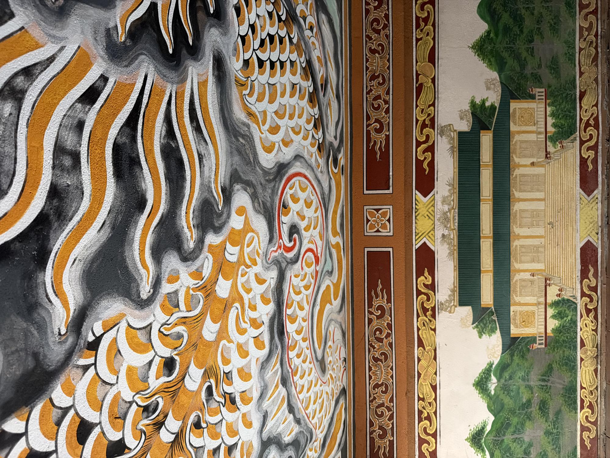 The image depicts a ceiling mural with intricate artwork, featuring a dragon motif rendered in shades of black, white, and gold. The dragon's scales are detailed with overlapping white and orange semi-circles, outlined in black. Swirling grey and white clouds, interspersed with wavy orange and white lines, surround the dragon. Below the ceiling mural is a section of ornate decorative molding in red and gold. Further down, a painted landscape is visible, showcasing a traditional East Asian architectural structure with a green roof, light-colored walls, and a wide staircase leading to its entrance. The building is nestled amongst lush green trees and rolling hills, painted in a muted, stylized manner. The lower edge of the image shows more decorative gold and red banding. This artwork is located in the Cua Dai Beach area of Hoi An, Vietnam, within a building that appears to be a temple or a traditional hall, possibly part of a resort or a private residence. There are no people or animals depicted in the image.