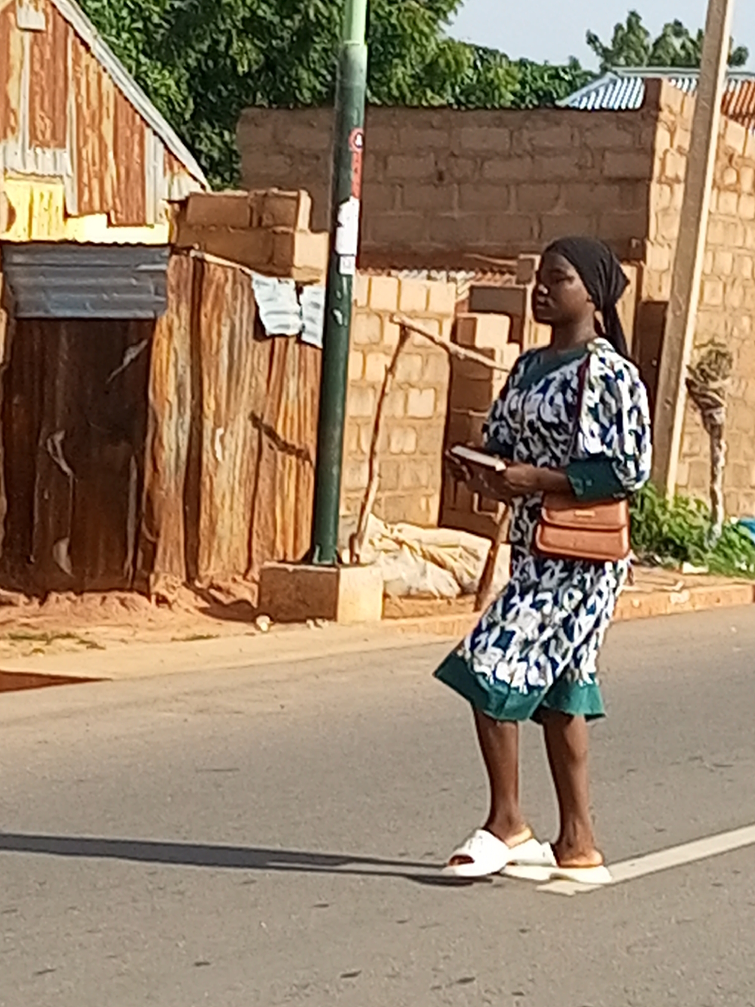 A woman in a black headscarf and a patterned blue and white dress with green trim is walking on a paved road in Tempure, Nigeria. She is carrying a brown handbag over her right shoulder and holding a book in her left hand. She is wearing white slip-on sandals. In the background, there is a building constructed with brick and corrugated metal. A green pole is visible on the right side of the image. The environment appears to be a sunny day with a shadow cast on the road.