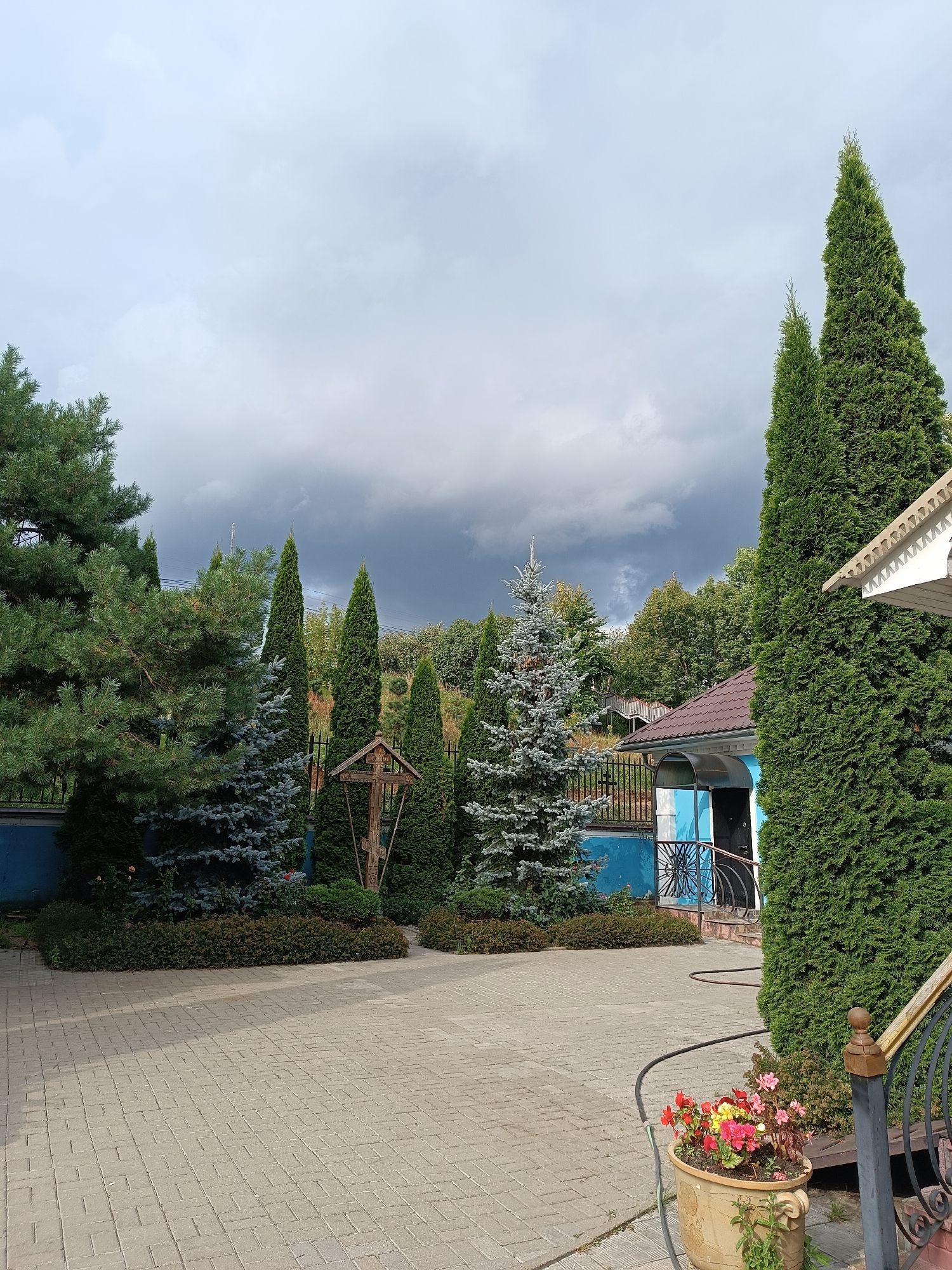 The image depicts an outdoor scene in Bykovo, Russia, likely a park or religious site, characterized by a paved courtyard, lush greenery, and a cloudy sky. In the foreground, a light-colored paved area, made of rectangular bricks laid in a uniform pattern, dominates the lower half of the image. To the right, a large, dense evergreen tree, possibly a thuja or cypress, stands tall, partially obscuring a building with a brown tiled roof and blue-and-white walls. A small covered entrance with a dark door is visible. Further to the left, a variety of evergreen trees, including pines and blue spruces, are arranged in groups, with a wooden cross-shaped structure placed among them. Behind the trees, a fence and a sloping hill covered in vegetation are visible. A pot of colorful flowers, with red, yellow, and pink blooms, sits on the paved ground near the right edge of the frame. The sky is overcast with dark, heavy clouds, suggesting imminent or recent rainfall. There are no people visible in the scene.
