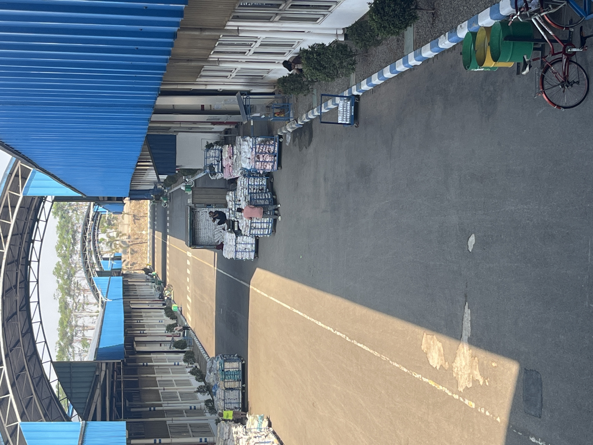 The image depicts an outdoor industrial area, likely a loading dock or distribution center. The scene is characterized by long, blue-roofed buildings with white walls and numerous windows. A paved road runs through the center, marked with a white line. Stacks of white sacks, some on wheeled carts, are visible throughout the area, suggesting the movement or storage of goods.
In the foreground, a red bicycle is parked next to three large cylindrical bins (two green, one yellow). Further down the road, several people are present. One individual in a yellow vest stands near a cart loaded with sacks. Another person, wearing a pink shirt, walks near a truck. Two individuals are seated near the building on the right.
The environment appears to be sunny with shadows cast across the road. Trees line the background behind the buildings. The location is identified as Pelang, Indonesia. The overall impression is one of an active, functional industrial space.