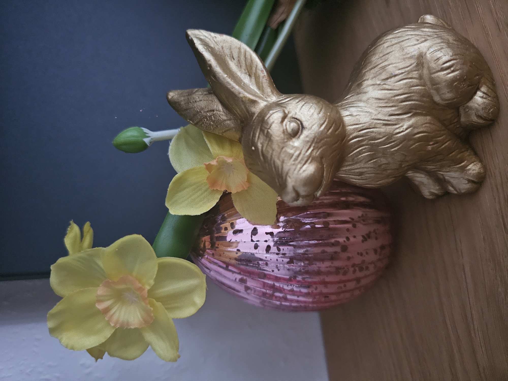 An arrangement of Easter-themed decorations sits on a wooden surface. In the foreground, a golden rabbit figurine is positioned facing left. To the left of the rabbit, a pink, ribbed, egg-shaped vase is visible. Emerging from behind the vase are several yellow daffodils with orange centers, attached to green stems. One daffodil is fully bloomed, while another is partially open, and a green bud is also present. The background is dark and out of focus. The lighting is soft, highlighting the textures and metallic sheen of the objects.
