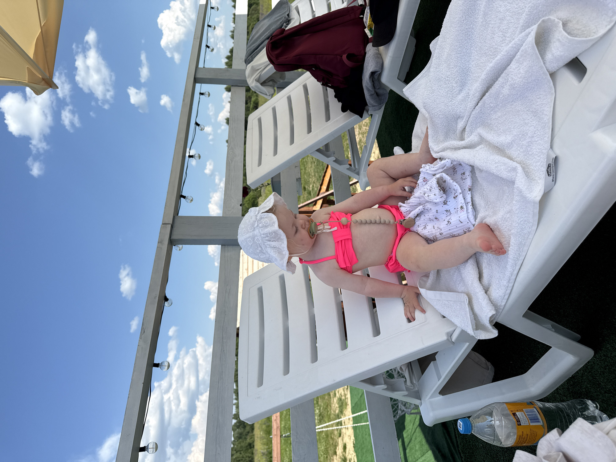 The media file shows a young child, likely a toddler, lying on a white plastic lounge chair. The child is wearing a pink bikini and a white sun hat, and has a pacifier with a beaded string in their mouth. They are covered partially by a white towel. Beside the child is a white cloth with a floral pattern. The lounge chair is situated outdoors, on what appears to be artificial turf. In the background, there is a wooden structure with string lights and a glimpse of green trees and a blue sky with white clouds. The overall impression is a warm, sunny day, suggesting a leisure activity like sunbathing or relaxing by a pool. The orientation of the image is rotated 90 degrees counter-clockwise. A plastic water bottle with a yellow label and blue cap is visible on the ground near the chair. The label on the water bottle is partially visible and appears to contain Cyrillic text. The location context is Колокша, Russia.