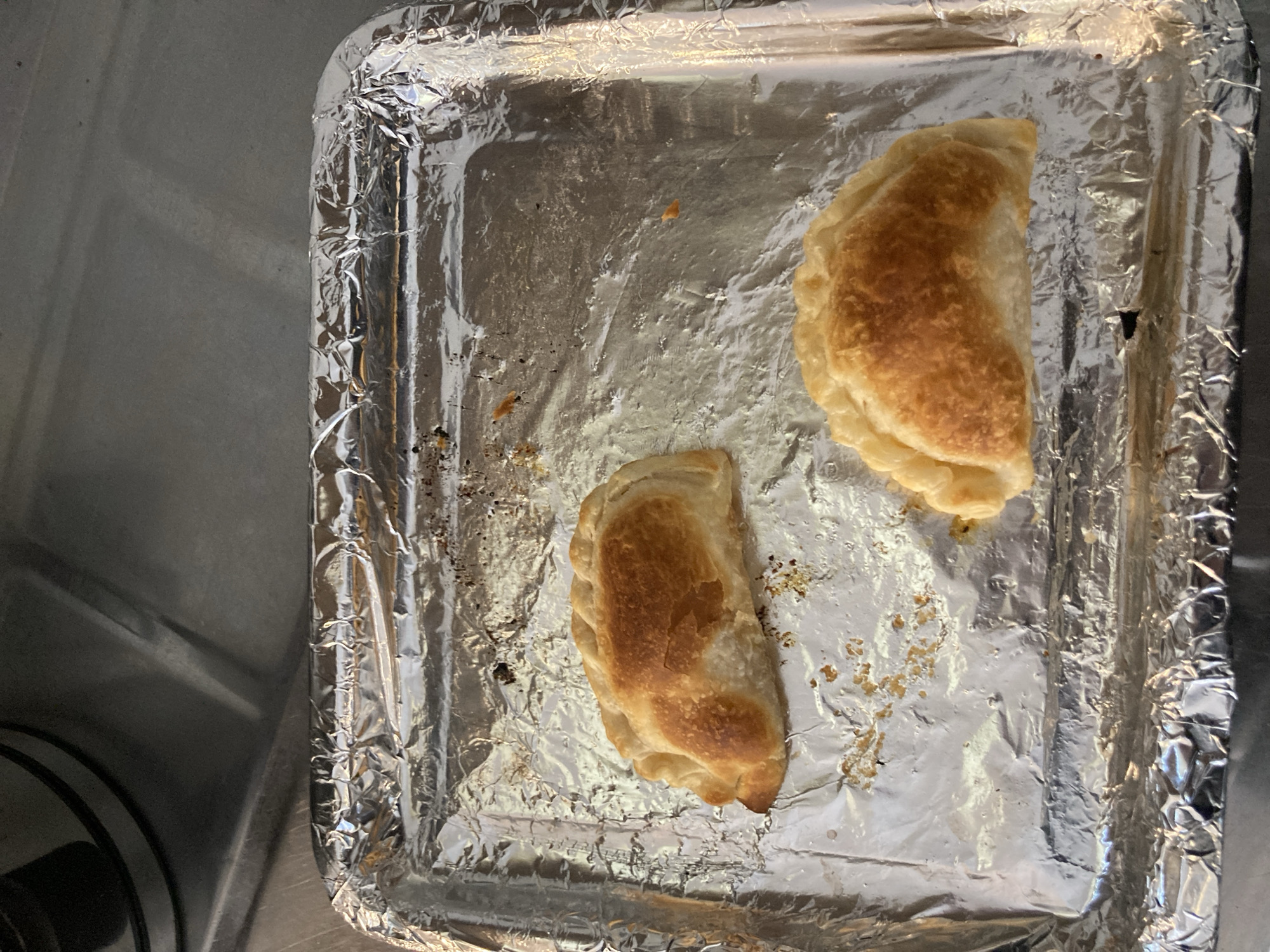 The image shows a metal baking sheet lined with aluminum foil, with two empanadas on it. The empanadas are golden brown and half-moon shaped. They are on a silver foil-lined baking sheet on top of what appears to be a kitchen sink. There is some debris from the food on the sheet. The location cannot be determined from the image, nor can a connection to Buenos Aires, Argentina.
