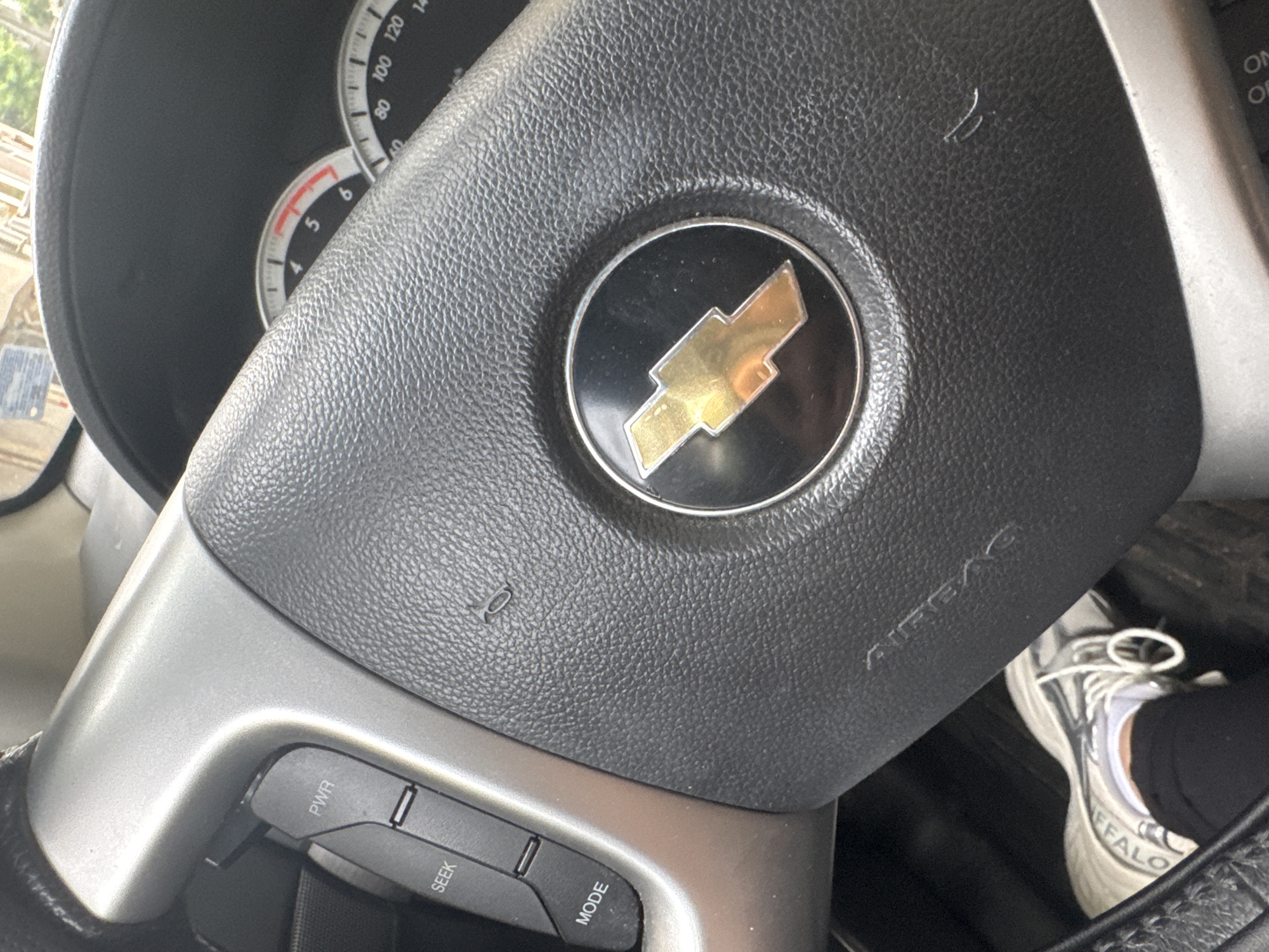 The image shows a close-up view of a car's steering wheel, focusing on the Chevrolet emblem and some of the dashboard controls. The steering wheel is made of black, textured material, with the Chevrolet bowtie logo prominently displayed in the center. The logo is a golden bowtie within a silver and black circle. To the left of the steering wheel, a portion of the car's instrument cluster is visible, showing a speedometer with markings up to 140 and a tachometer with markings up to 6. Below the steering wheel, on a silver-colored console, there are several buttons labeled "PWR," "SEEK," and "MODE." The overall scene is inside a car, likely during the daytime given the lighting. The image is taken from the driver's perspective. There are no people clearly visible, but a shoe is partially visible in the lower right corner, suggesting a person is present. The setting is implied to be a car interior. The visible text includes "PWR," "SEEK," "MODE," "AIRBAG," and numbers on the speedometer and tachometer. There are no specific location cues to suggest Plovdiv, Bulgaria, beyond the prompt's context.