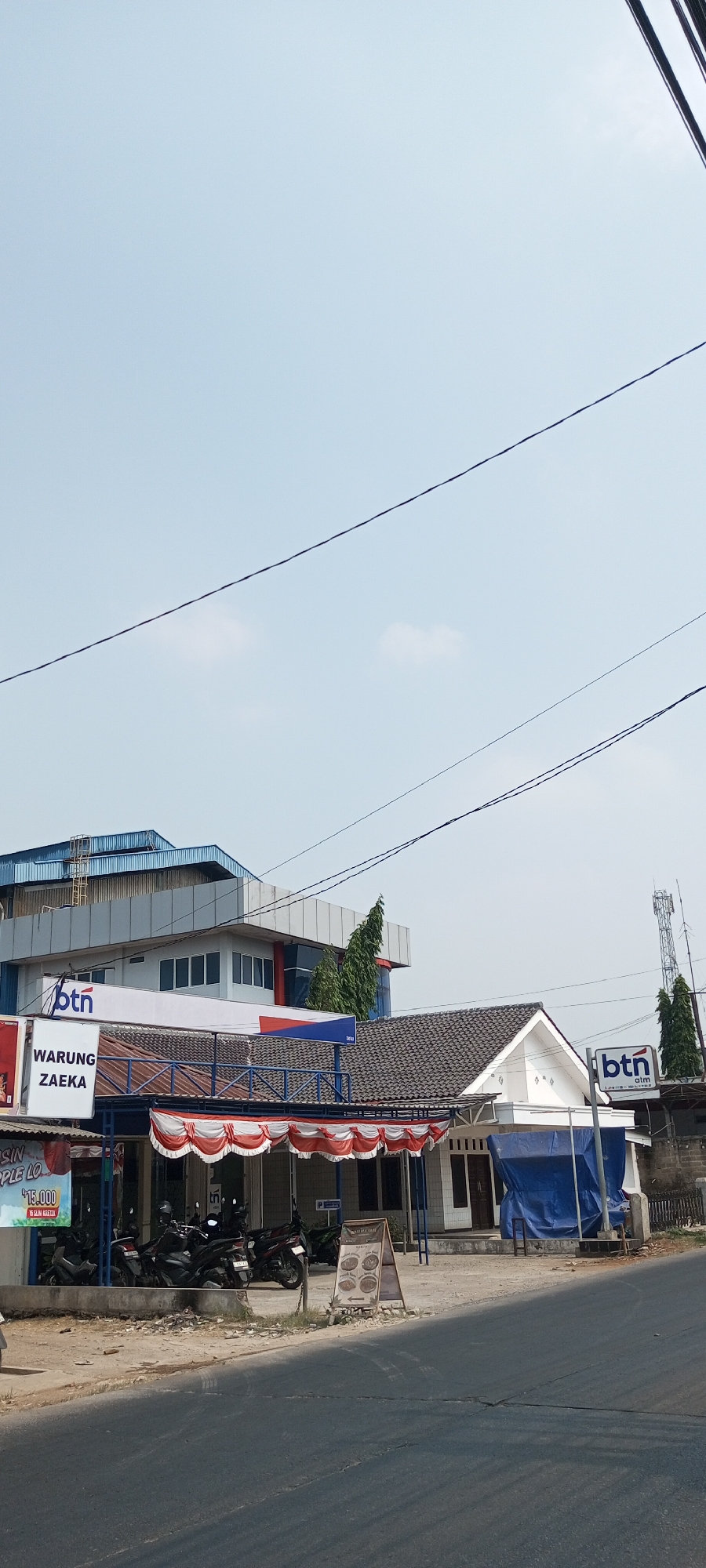 The image depicts a street scene in Setu, Indonesia. A modern, multi-story building with blue and gray accents stands on the left, featuring the "btn" logo. In front of this building, a warung, or small food stall, named "WARUNG ZAEKA" is visible, adorned with red and white decorative bunting. Several motorcycles are parked under a covered area in front of the warung. To the right of the warung, a smaller white building with a tiled roof is partially covered by a large blue tarp, with another "btn" logo displayed. A cell tower is visible in the background. The foreground shows a paved road with clear lane markings. The sky is clear with some scattered clouds.